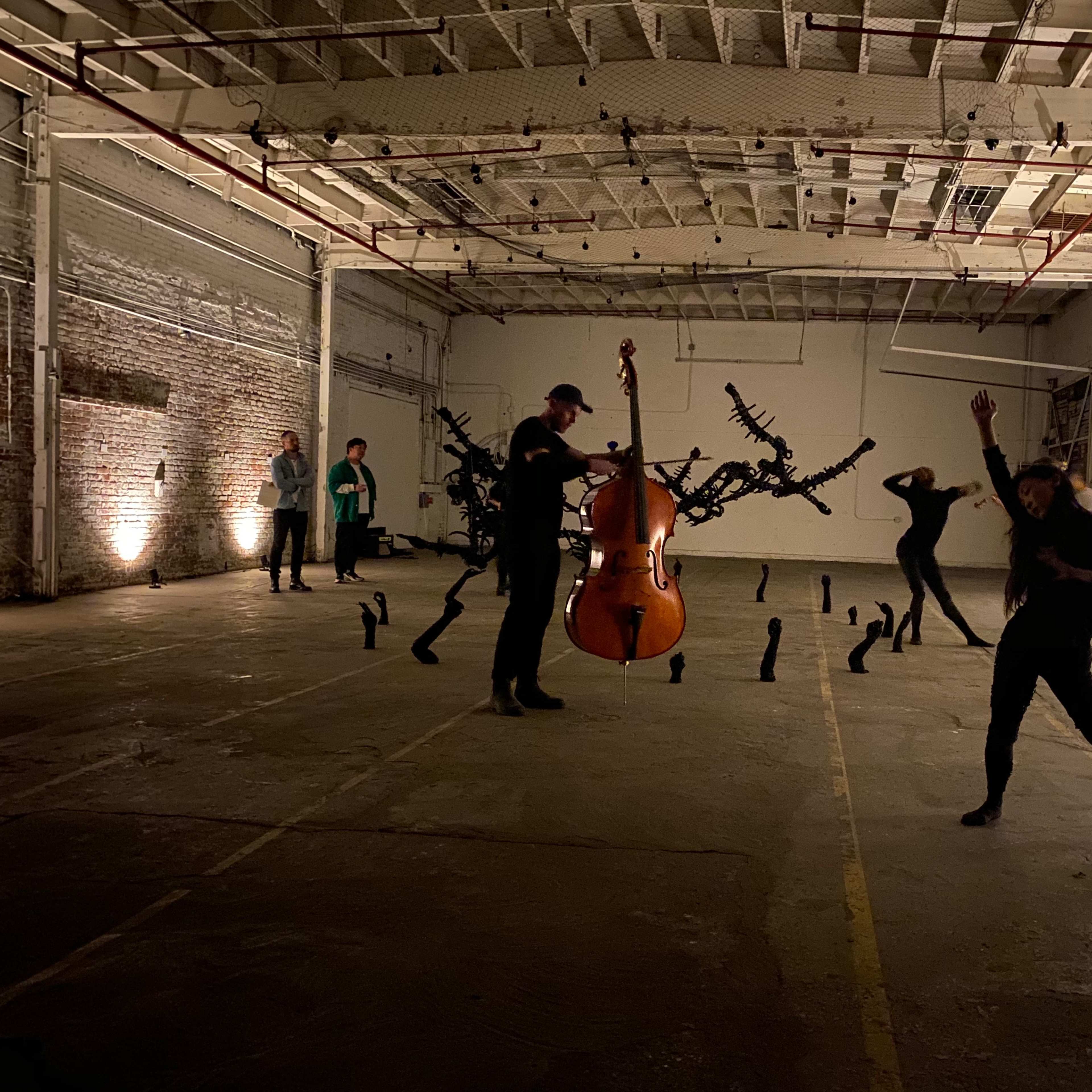 A contemporary art performance in an industrial space, featuring a cellist and dancers interacting with sculptural elements on the ground.