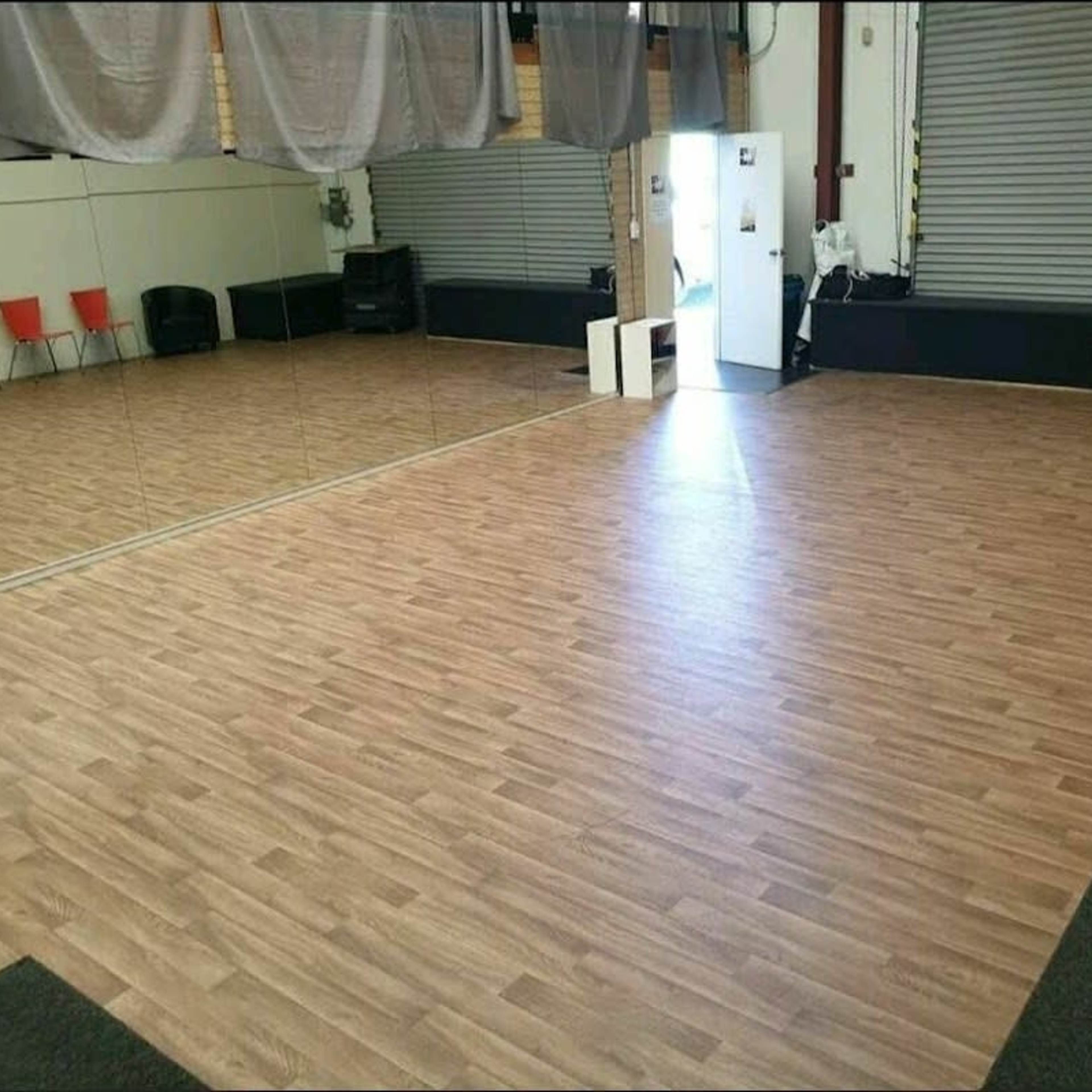 A spacious dance studio with wooden flooring, mirrors on one wall, and a few red chairs along the side.
