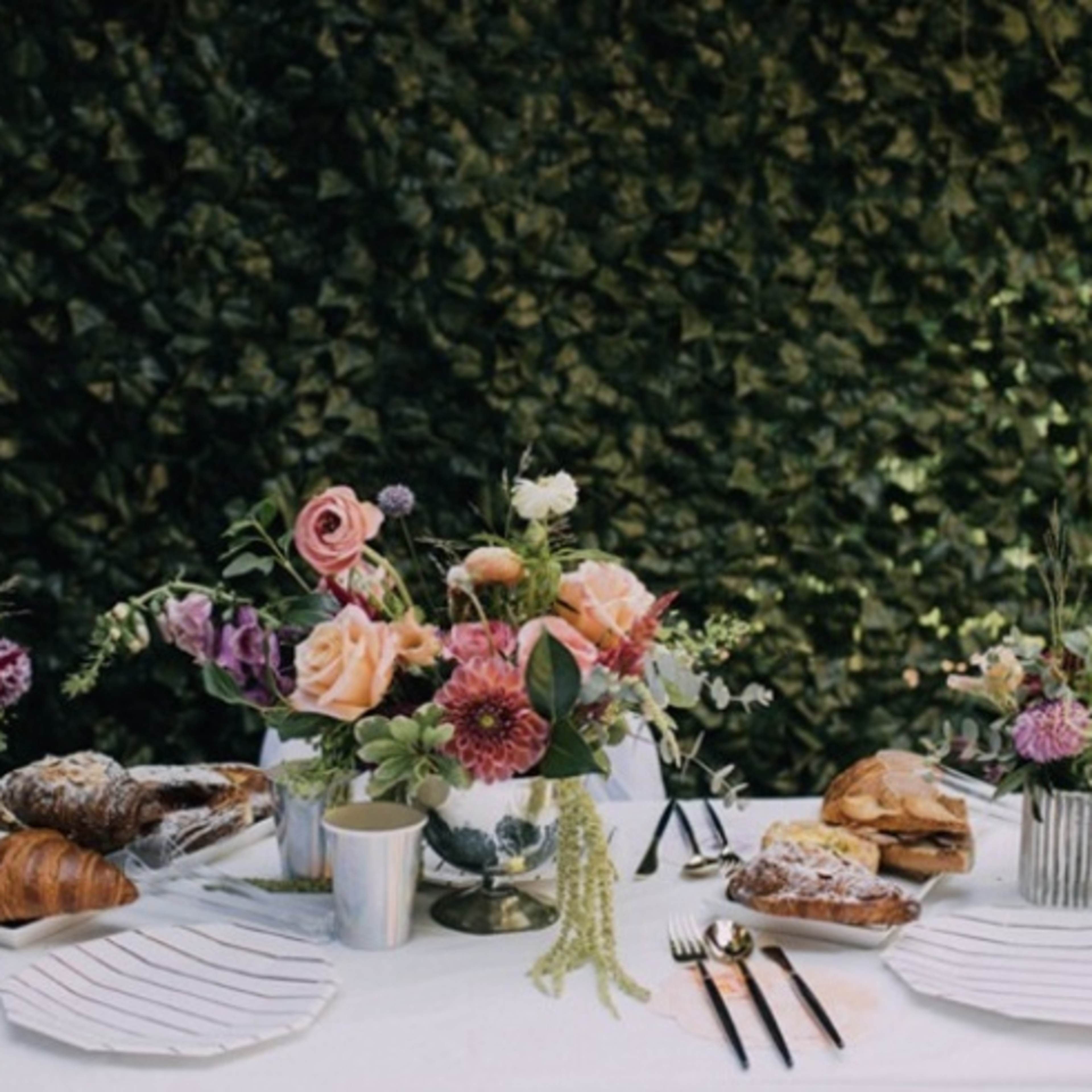 A table is set with flowers, croissants, and plates against a green foliage backdrop.