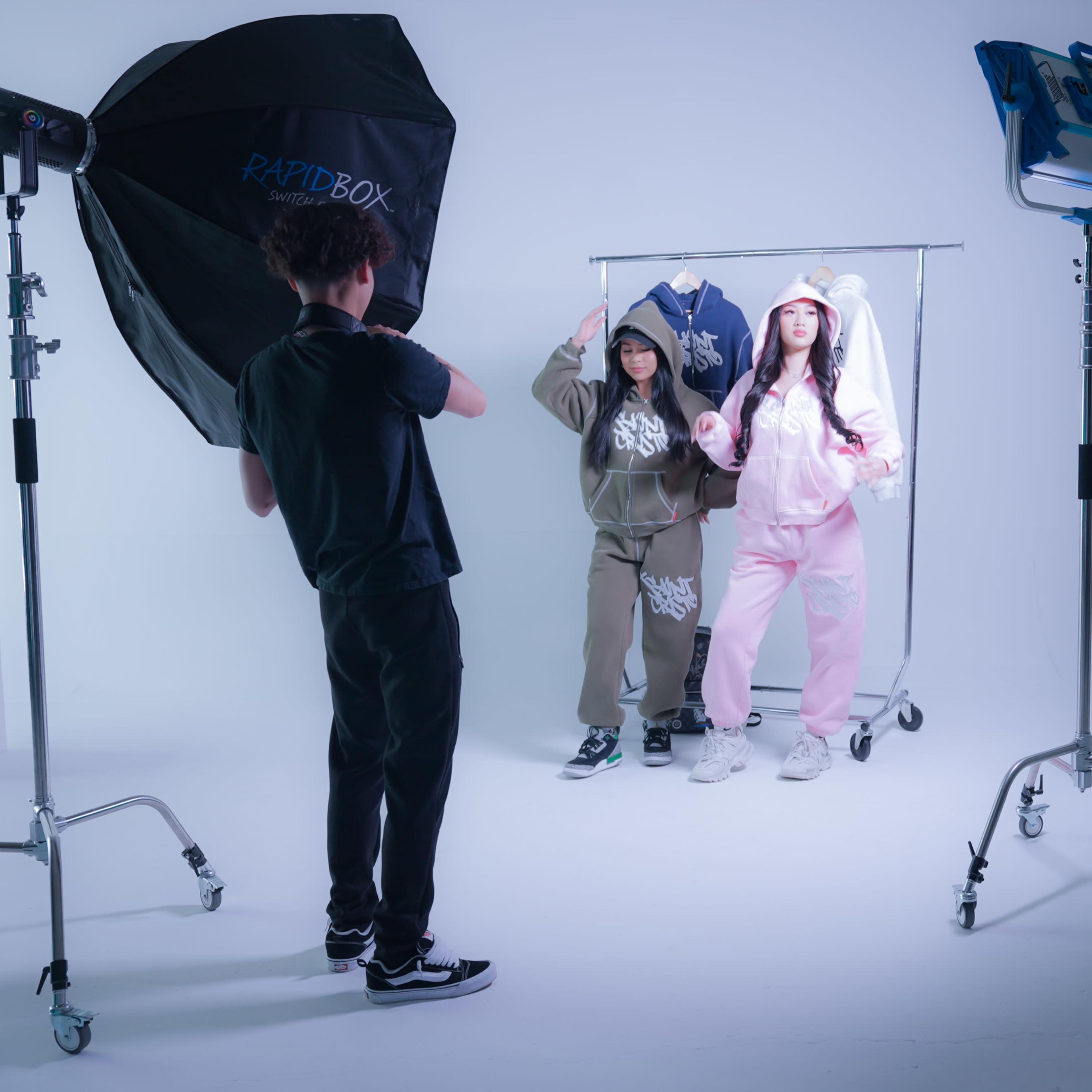 Two models, one in olive green and the other in pink, pose in front of a clothing rack in a photo studio while a photographer sets up lighting equipment.
