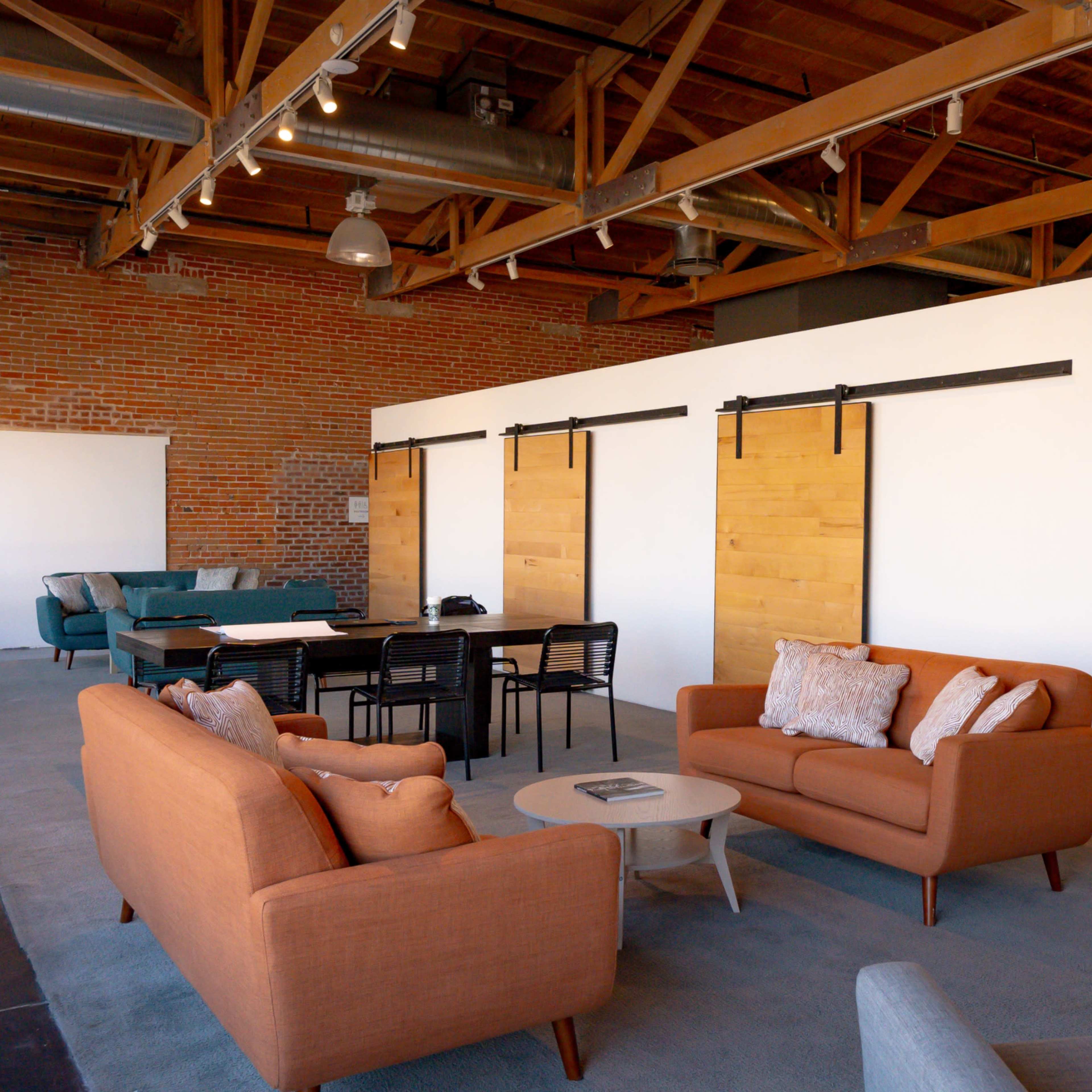 A modern, open-concept office space featuring a seating area with orange and gray sofas, a central table with black chairs, and sliding wooden barn doors against a brick wall.