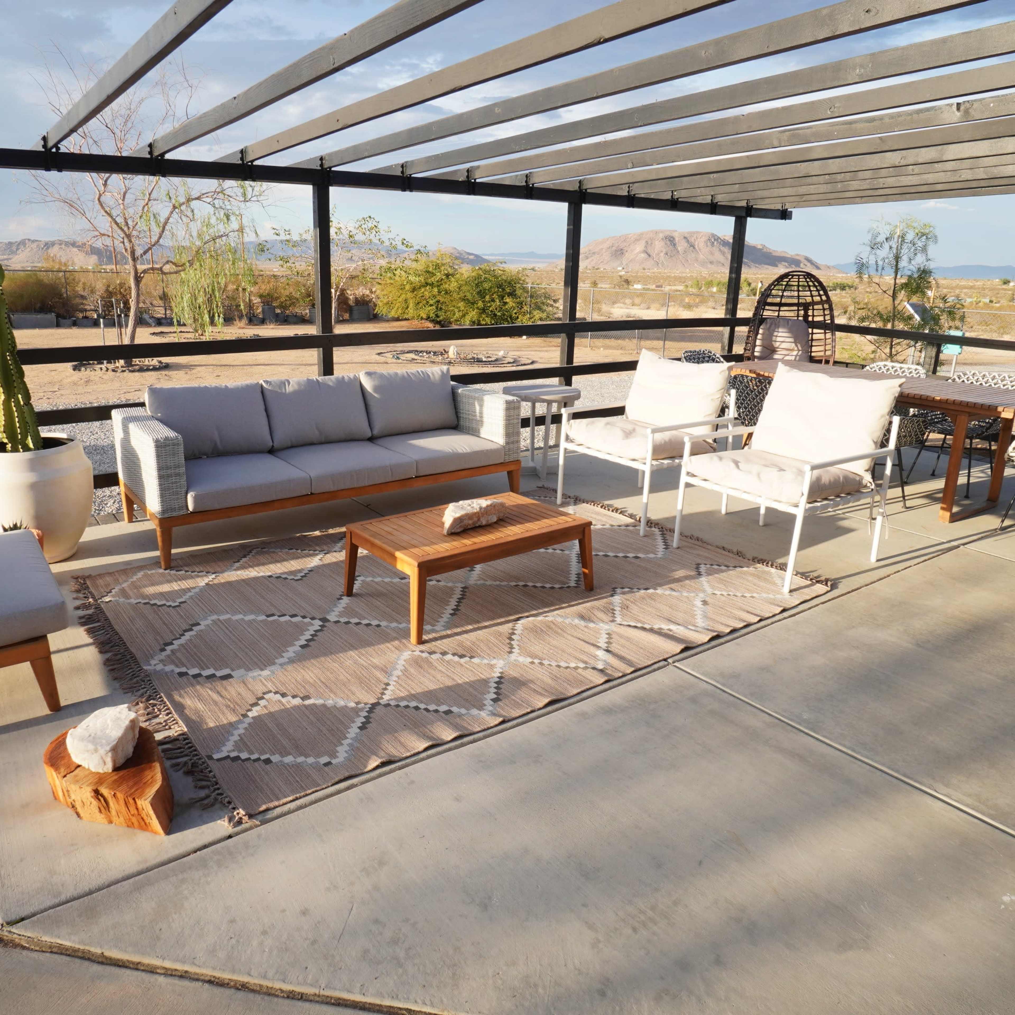 A covered outdoor seating area with modern furniture, including a sofa, chairs, and a wooden coffee table on a concrete surface, set against a desert landscape.