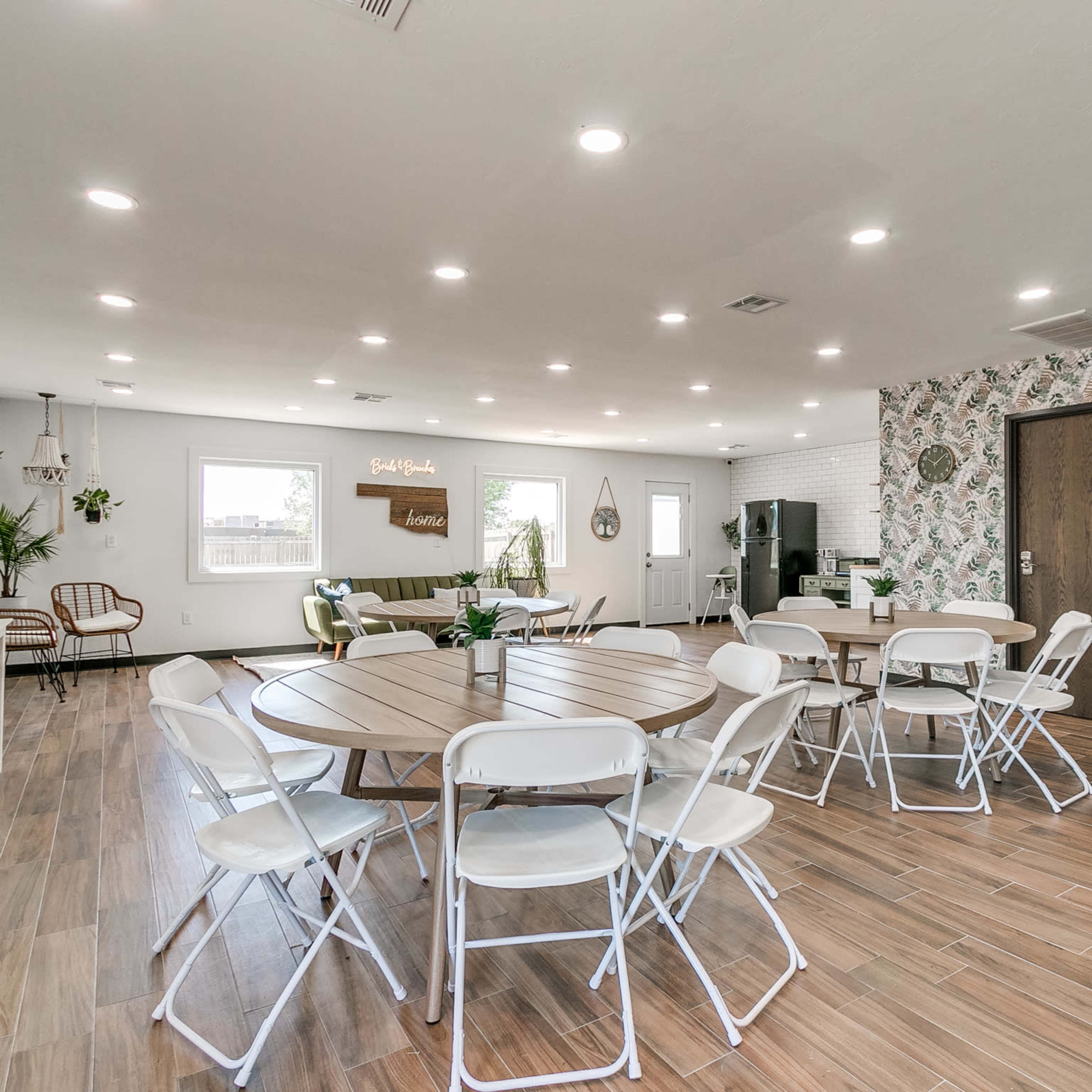 A spacious interior featuring multiple round tables with white chairs, a green sofa, and a modern kitchen area against a patterned wall.