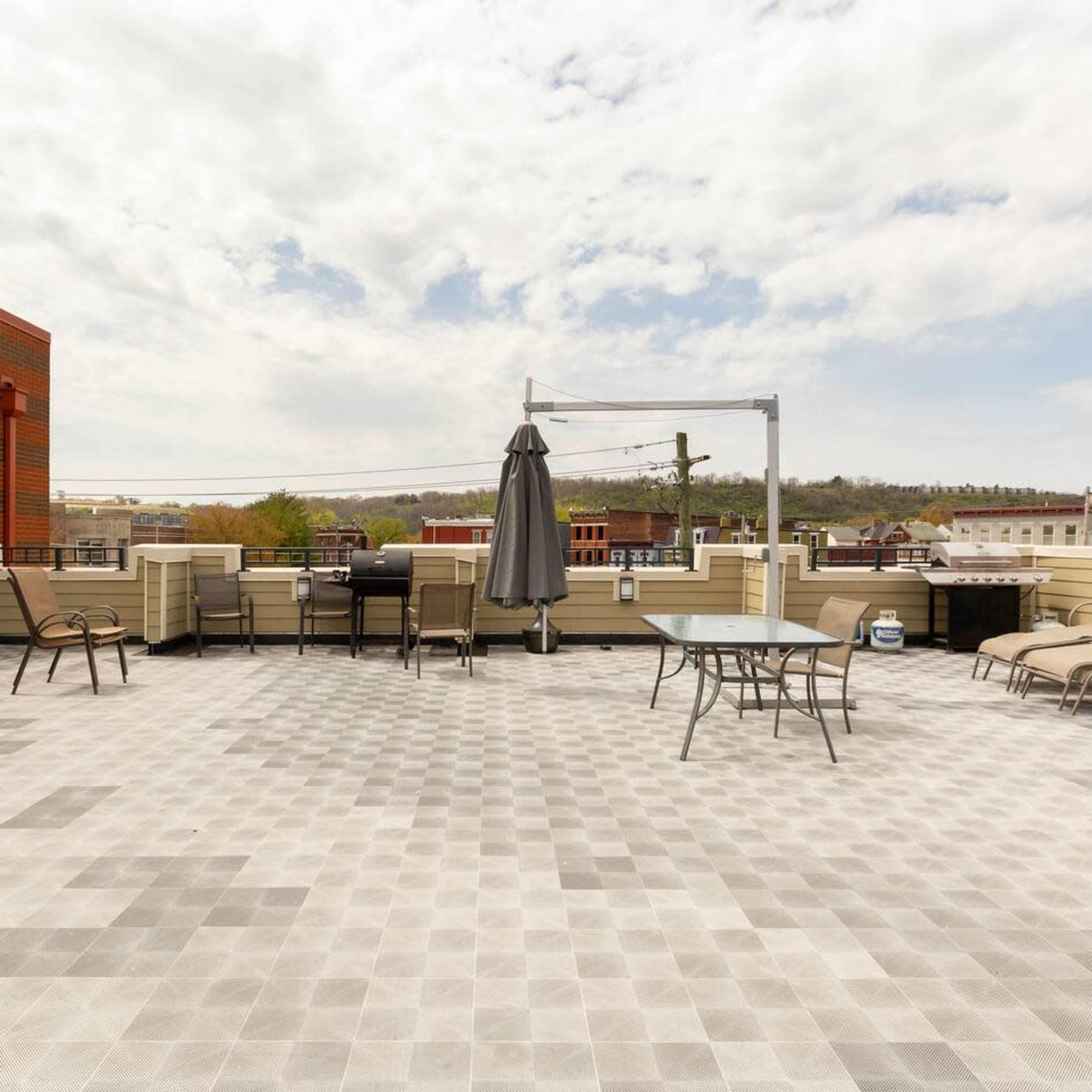A spacious rooftop terrace with chairs, tables, and a large umbrella against a backdrop of buildings and a cloudy sky.