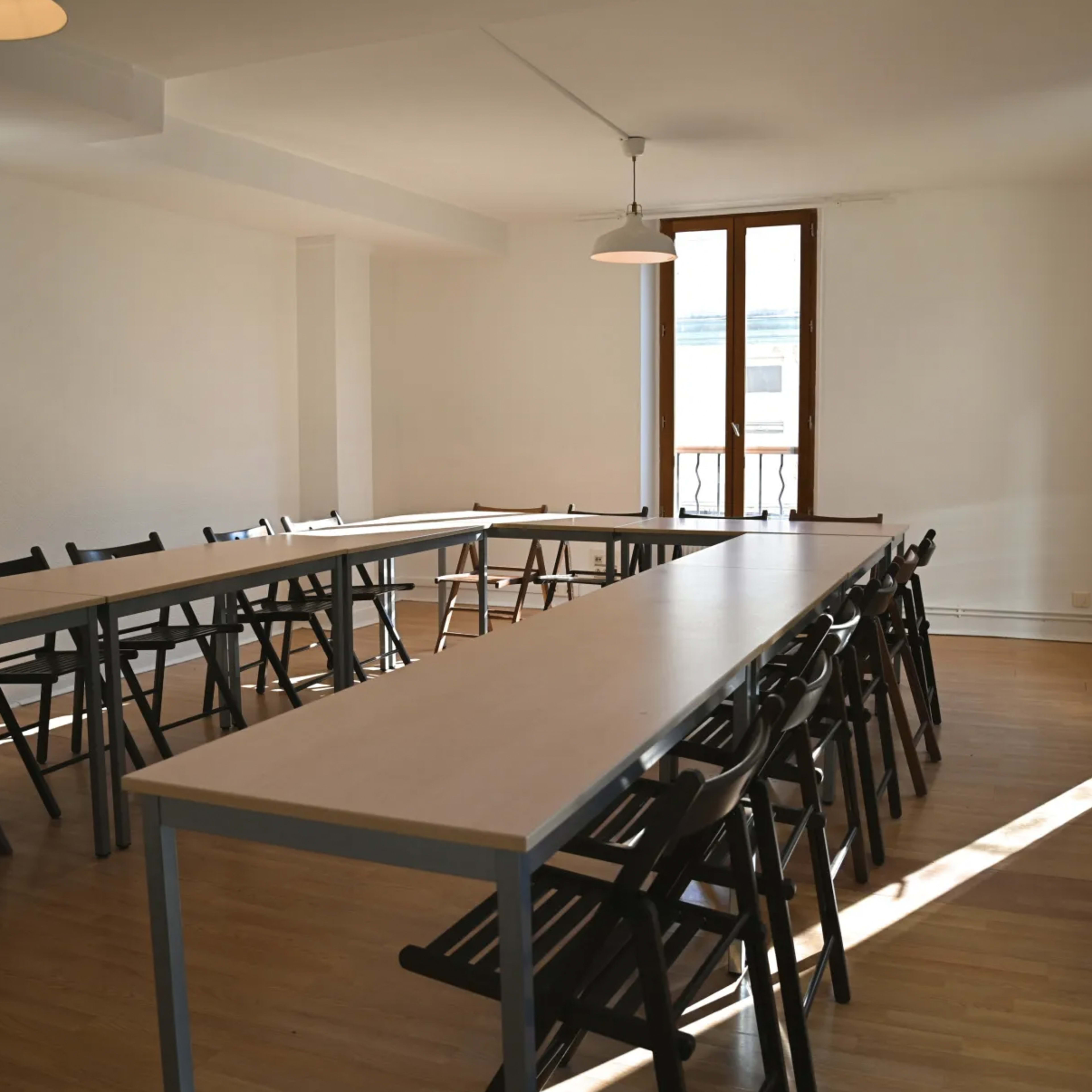 A spacious meeting room features several long tables arranged in a U-shape with black chairs around them and large windows allowing natural light.