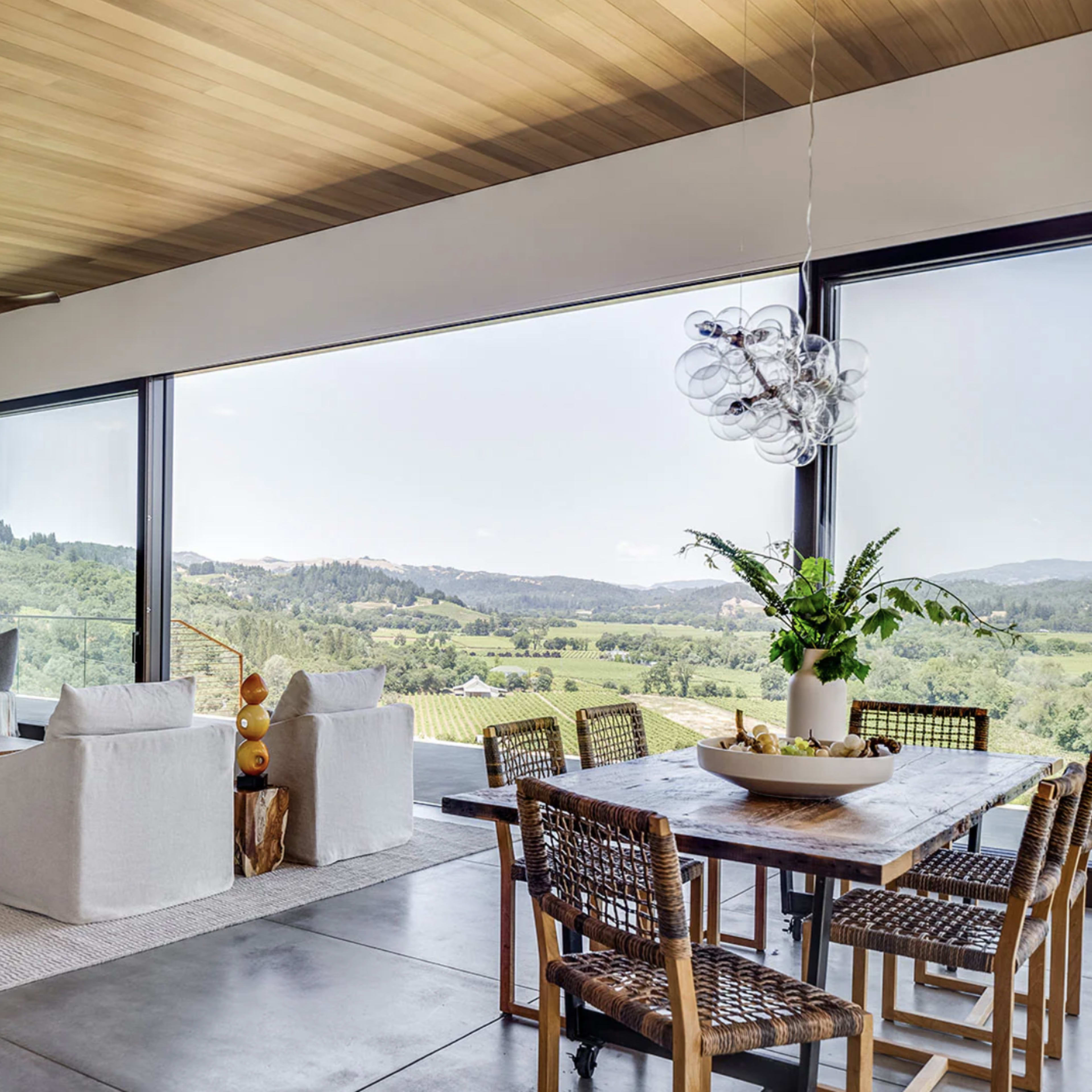 A modern living room features large windows with a scenic view, comfortable seating, and a dining area with a wooden table surrounded by woven chairs.