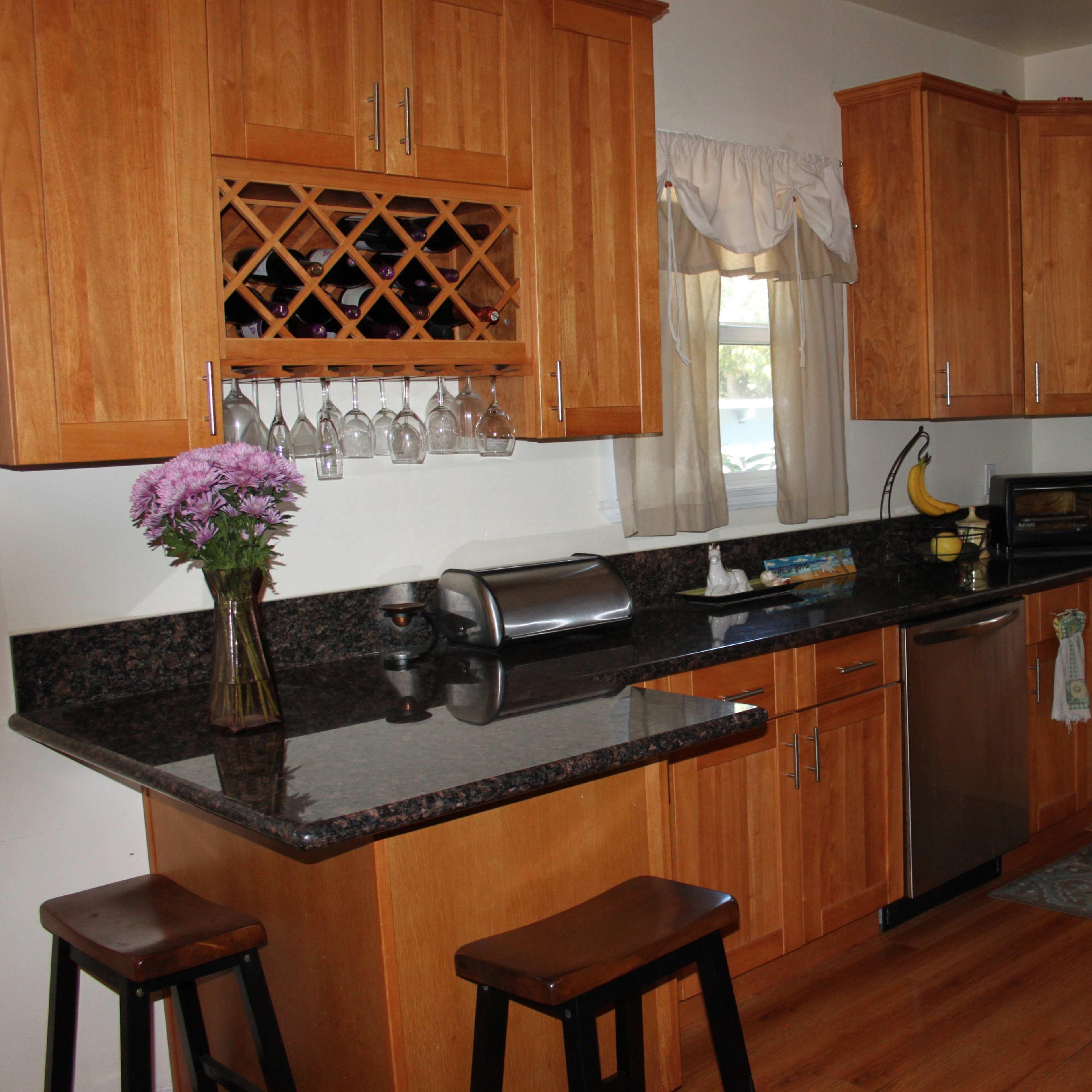 A kitchen with wooden cabinetry, a granite countertop, a wine rack, and a small dining area with two stools.