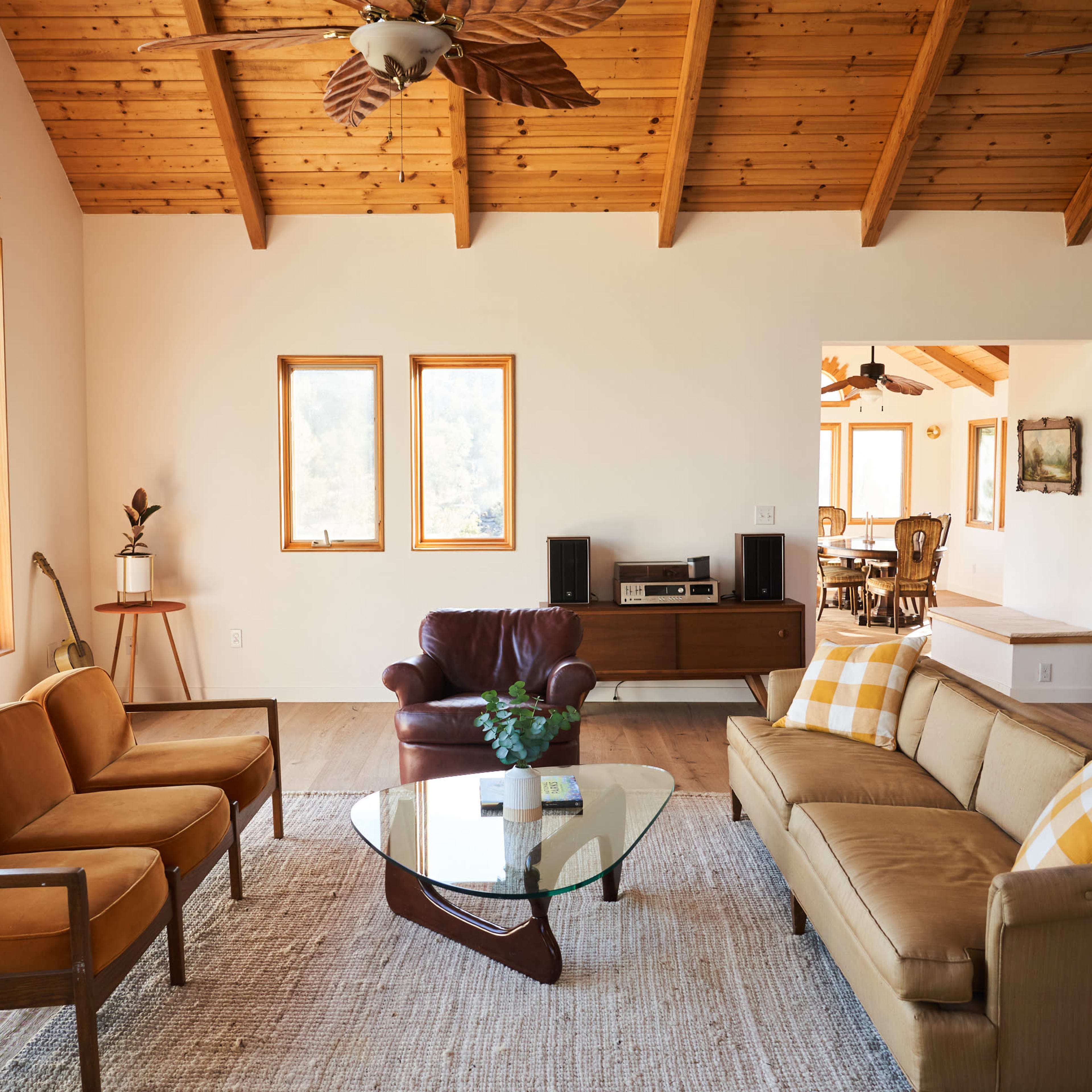 The living room features a combination of brown and beige furniture arranged around a glass coffee table under a wooden ceiling with exposed beams.