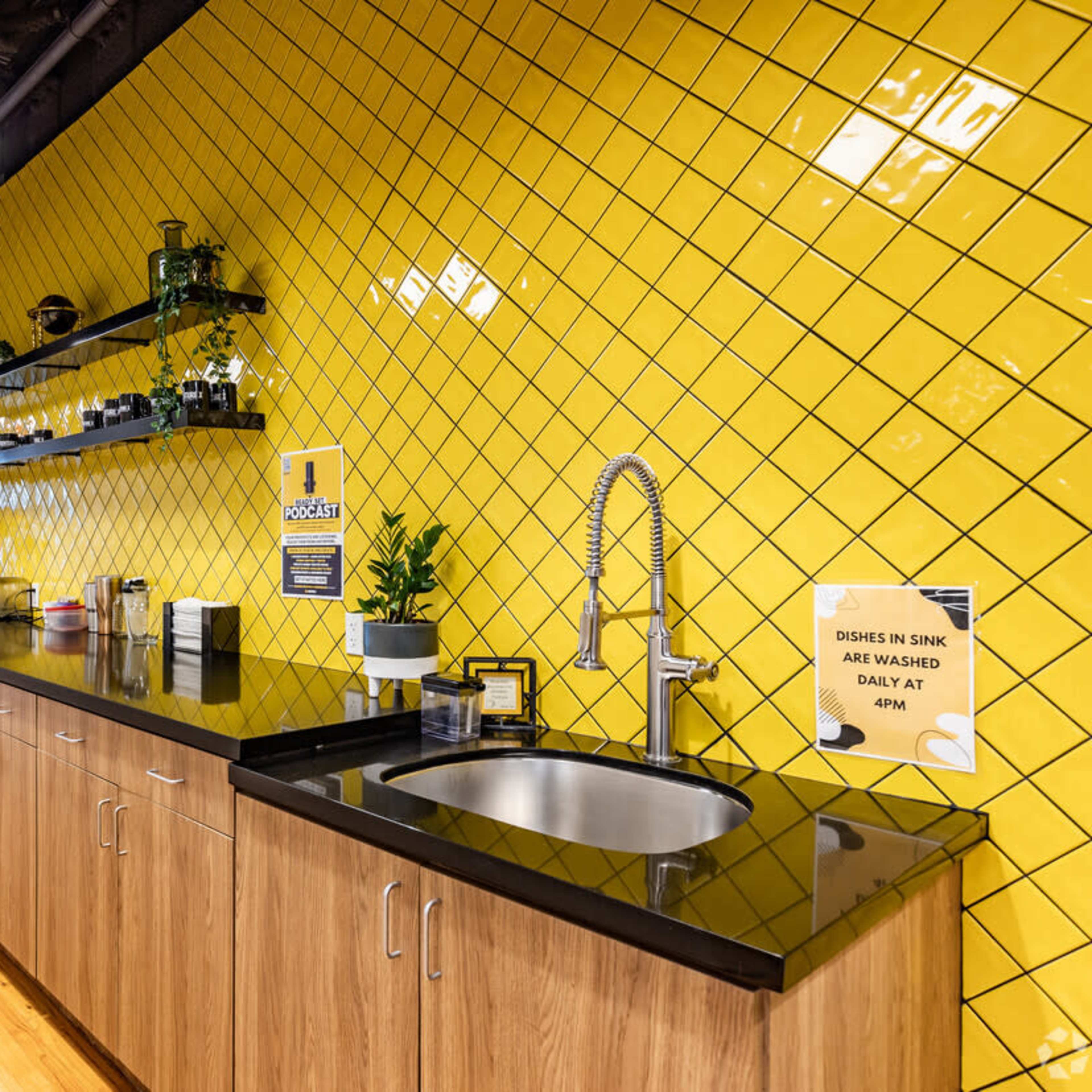 A modern kitchen area with a yellow tiled wall, black countertops, wooden cabinets, and various kitchen appliances.