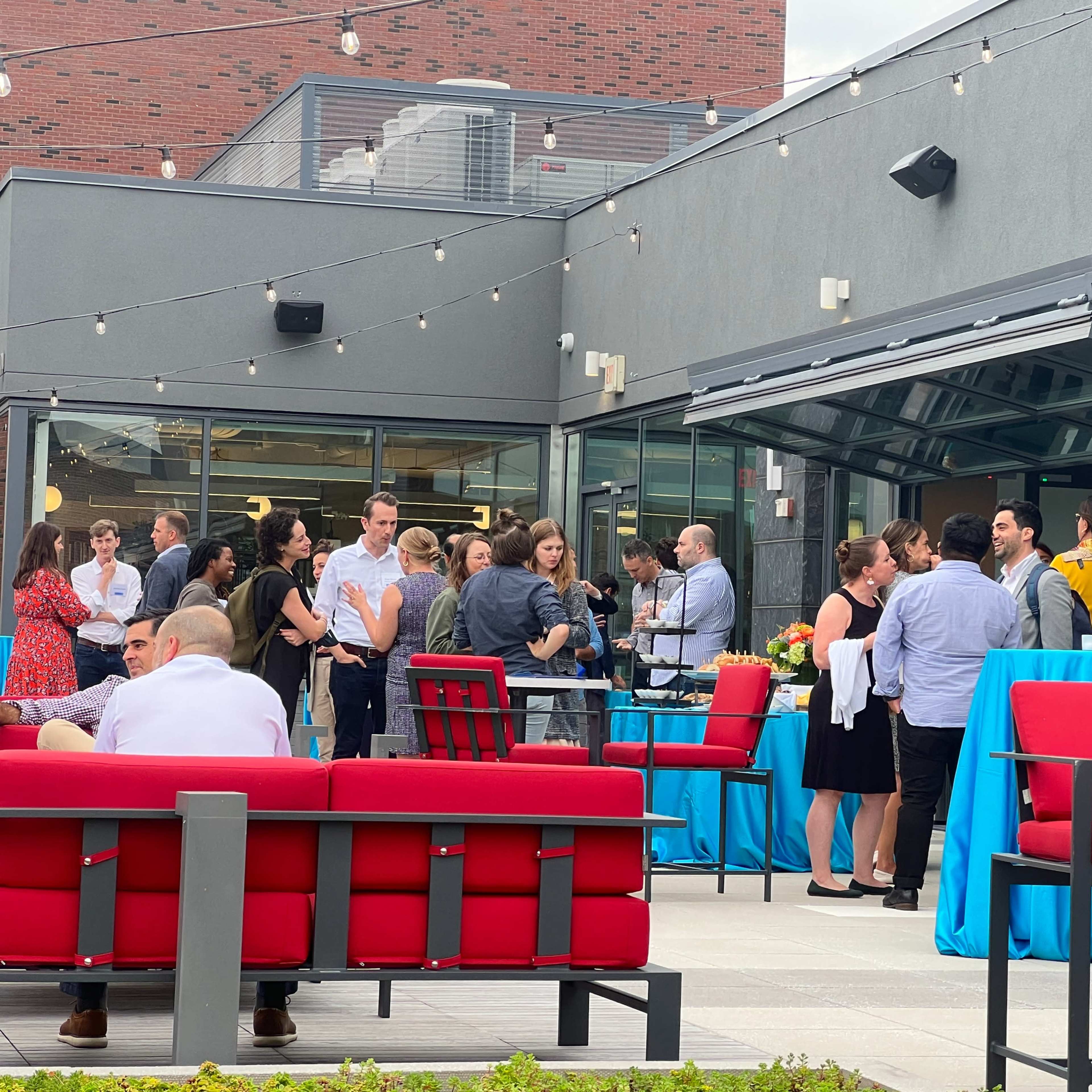 A group of people is gathered at an outdoor event on a rooftop, engaging in conversation around tables adorned with turquoise tablecloths.