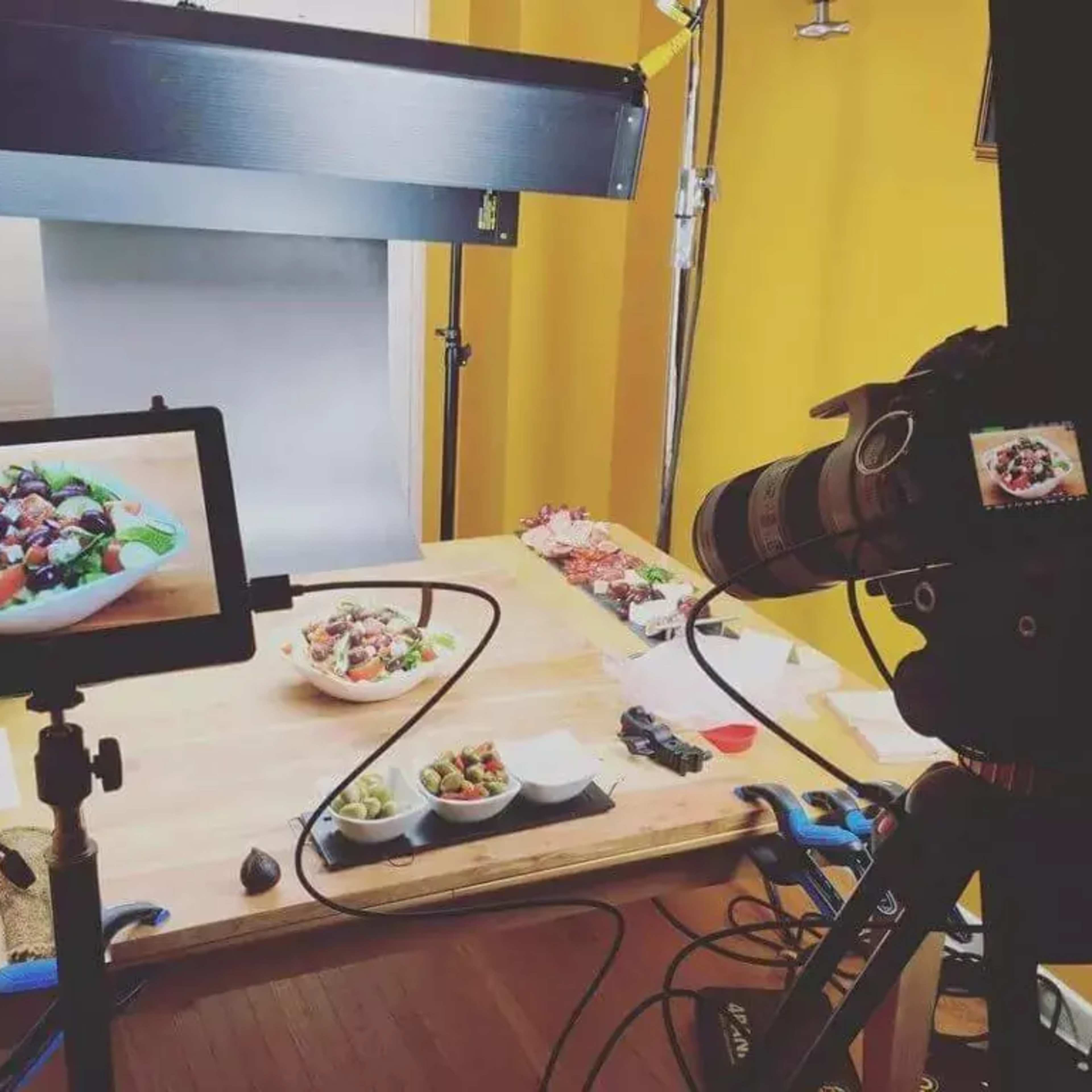 A food photography setup with a camera on a tripod, a monitor displaying a bowl of salad, and several dishes arranged on a wooden surface.