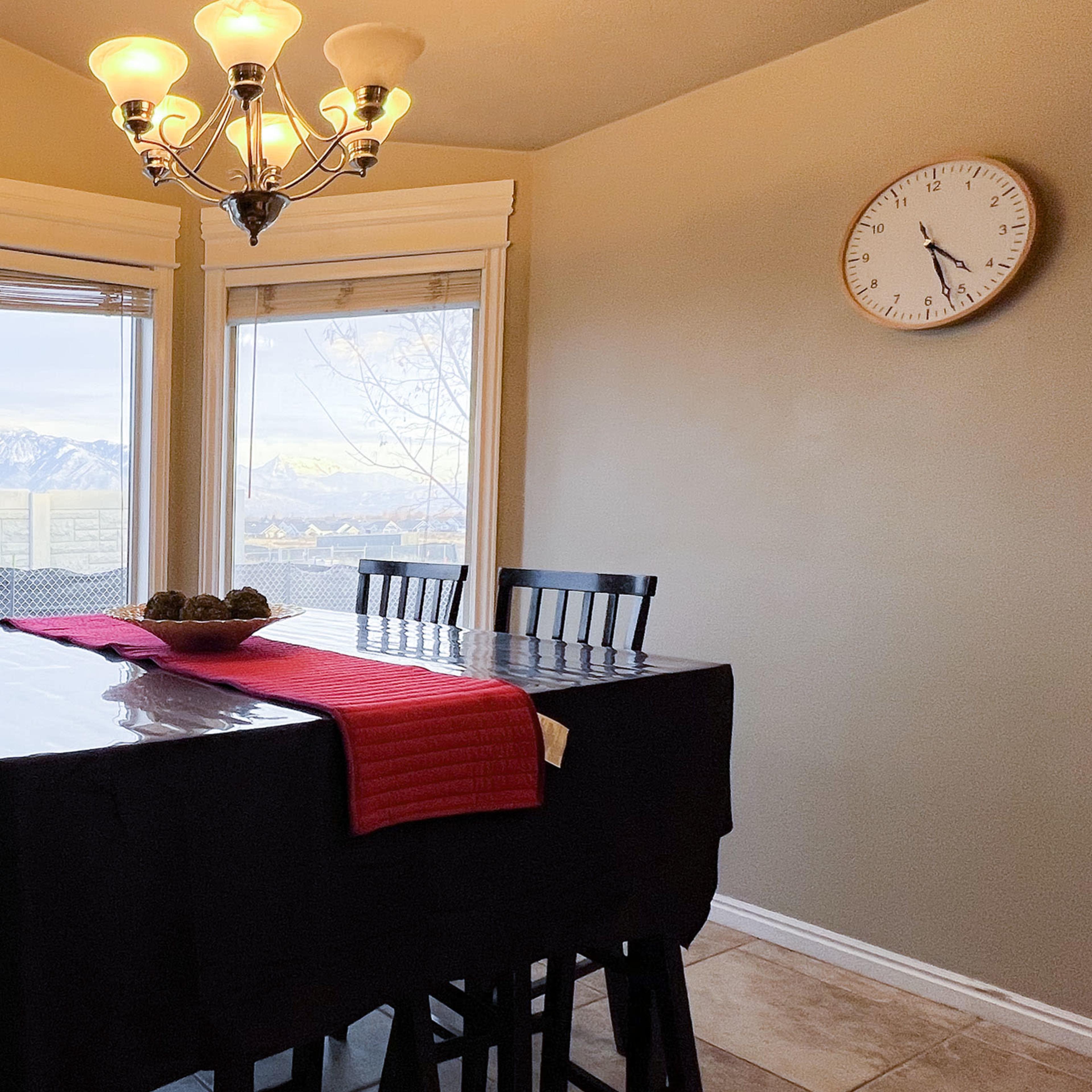 A dining area features a black table with a red runner surrounded by chairs, large windows showcasing a mountain view, and a chandelier above.