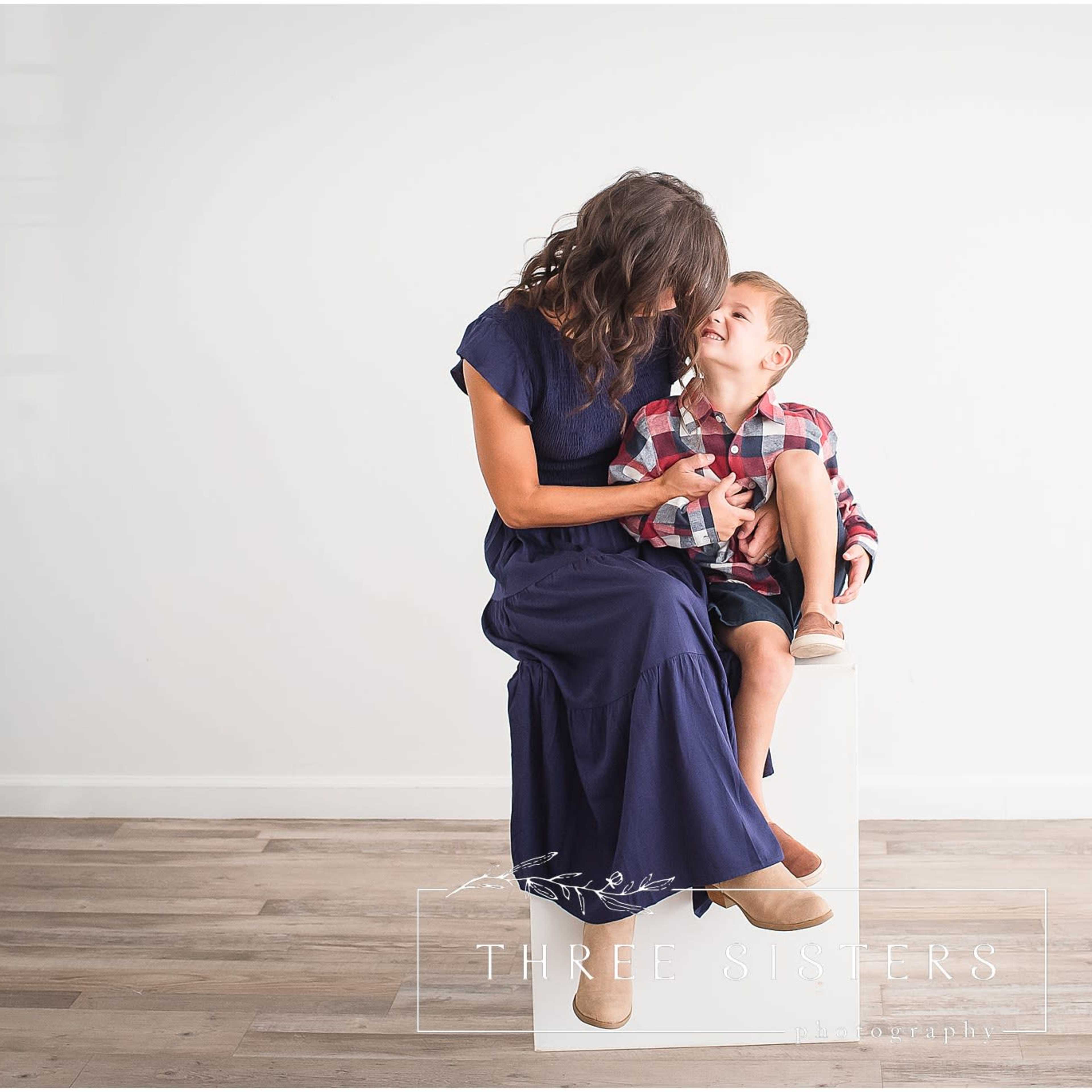 A woman in a navy dress sits on a pedestal while playfully interacting with a young boy wearing a plaid shirt and shorts.