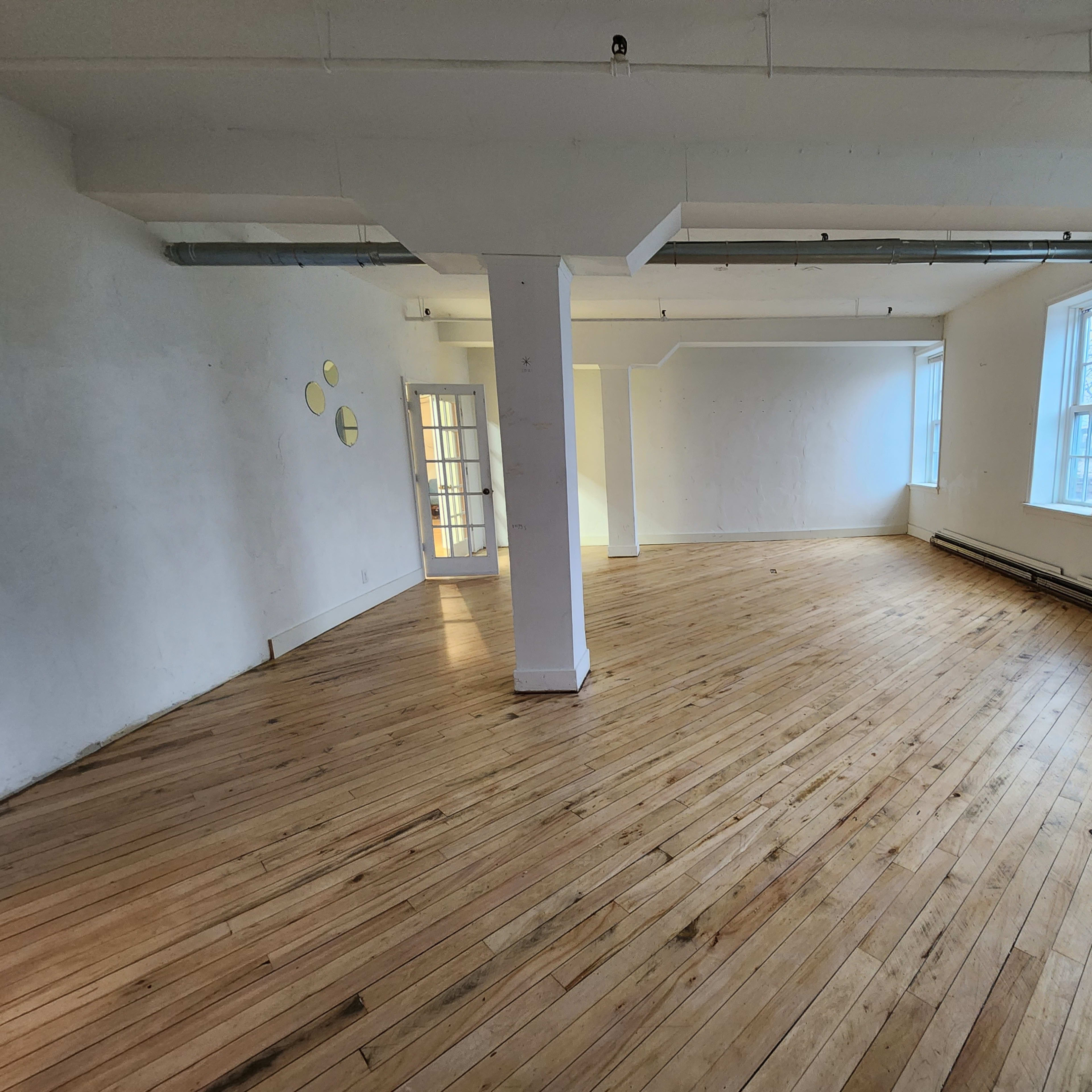 A spacious, empty room with wooden flooring, two windows, and a column in the center.