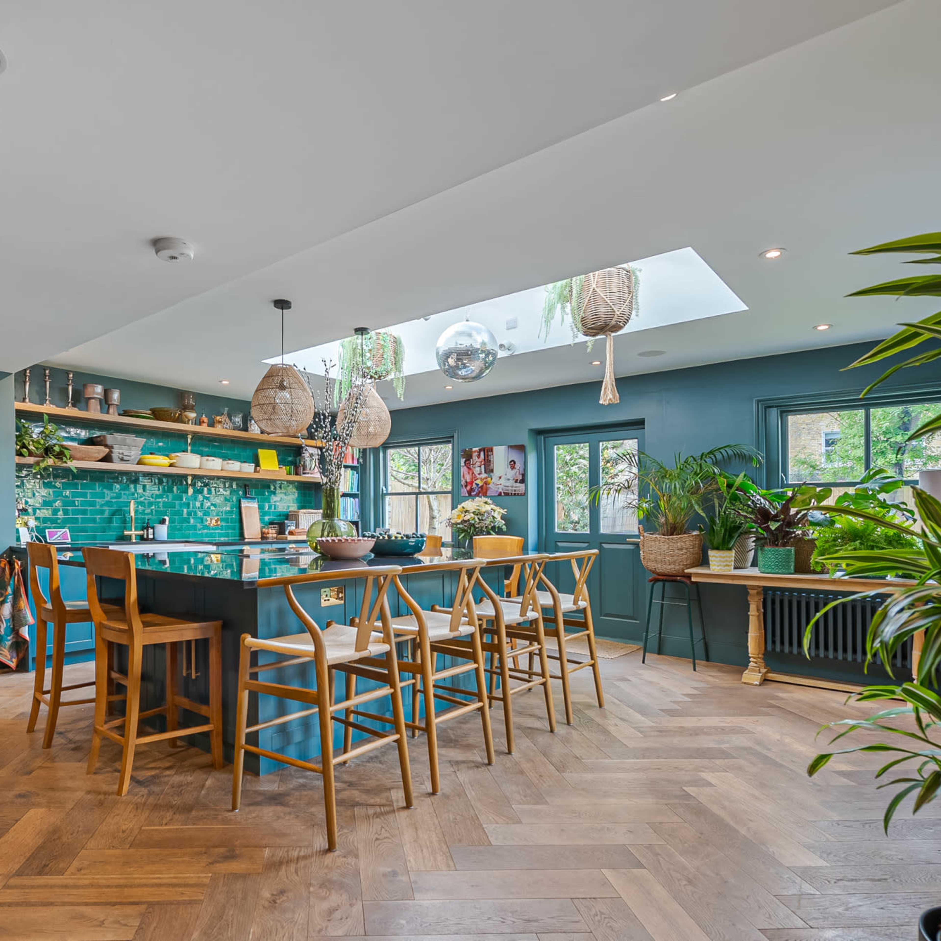 A modern kitchen with a large island, teal cabinetry, and a skylight, surrounded by plants and wooden flooring.
