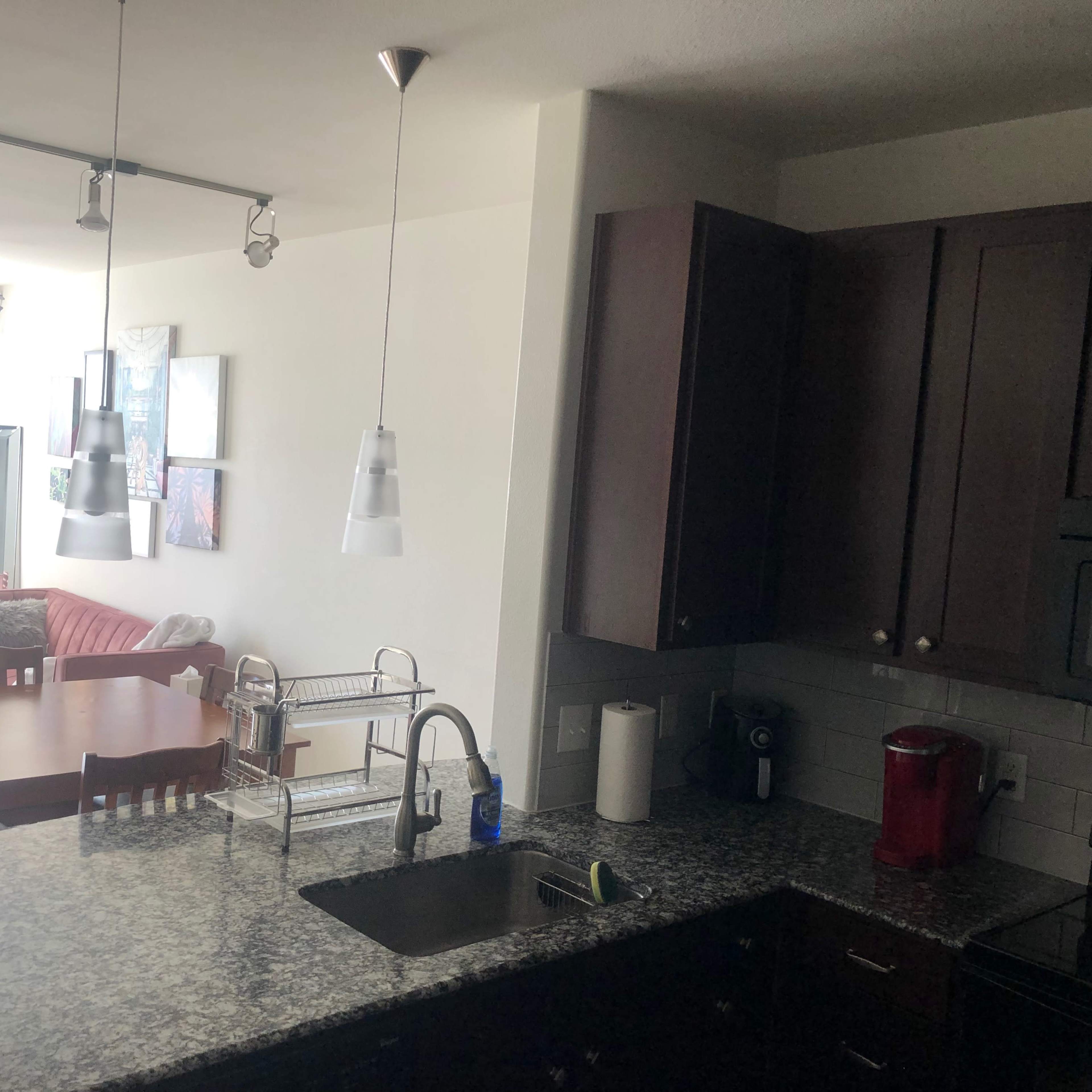 A kitchen with dark cabinetry and a granite countertop, connected to a living area featuring a dining table and a sofa.