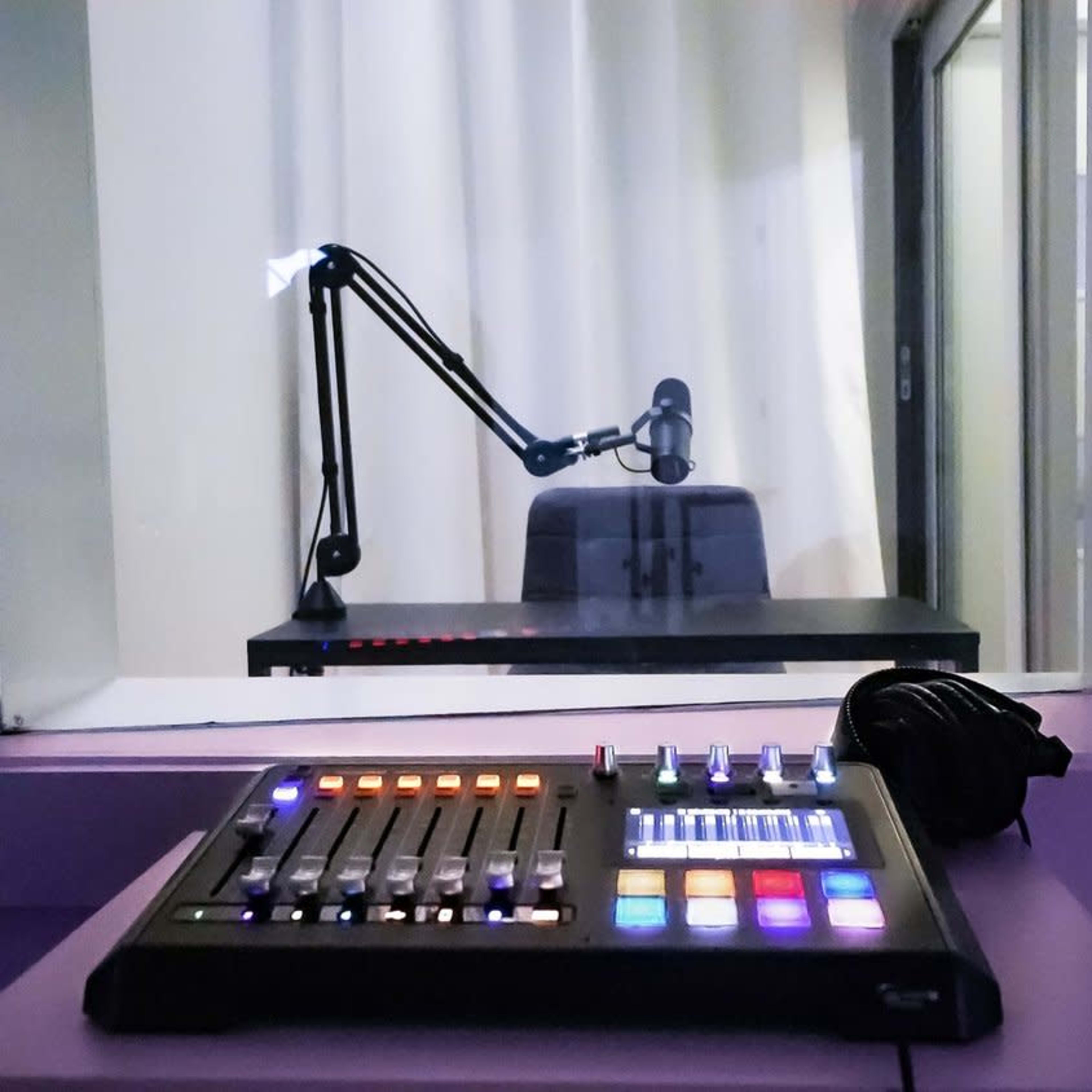 A control panel with various buttons and lights sits on a desk in the foreground, while a microphone arm and empty chair are visible through a glass window in the background.