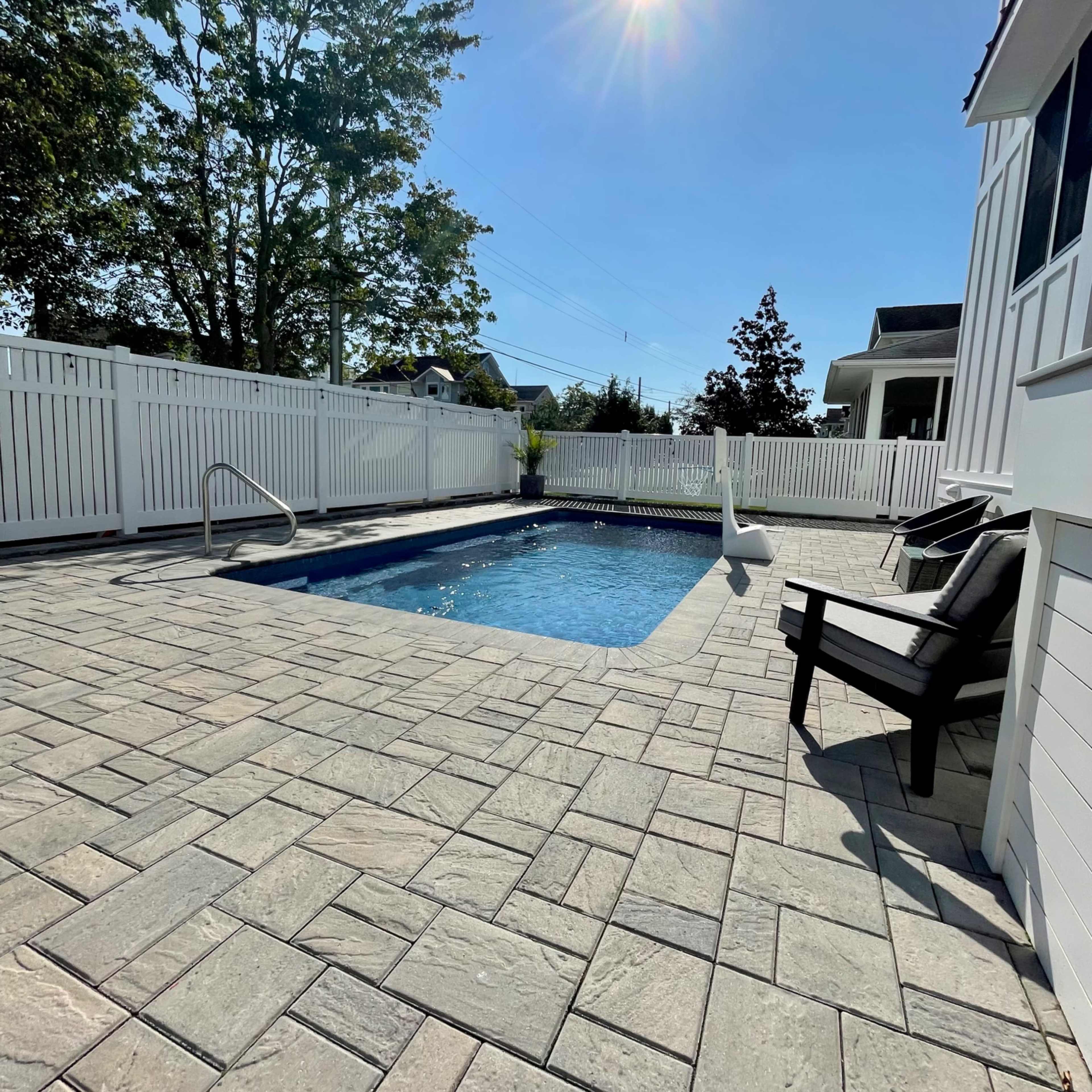A residential backyard with a rectangular swimming pool surrounded by a paved patio, lawn chairs, and a white fence under clear blue skies.