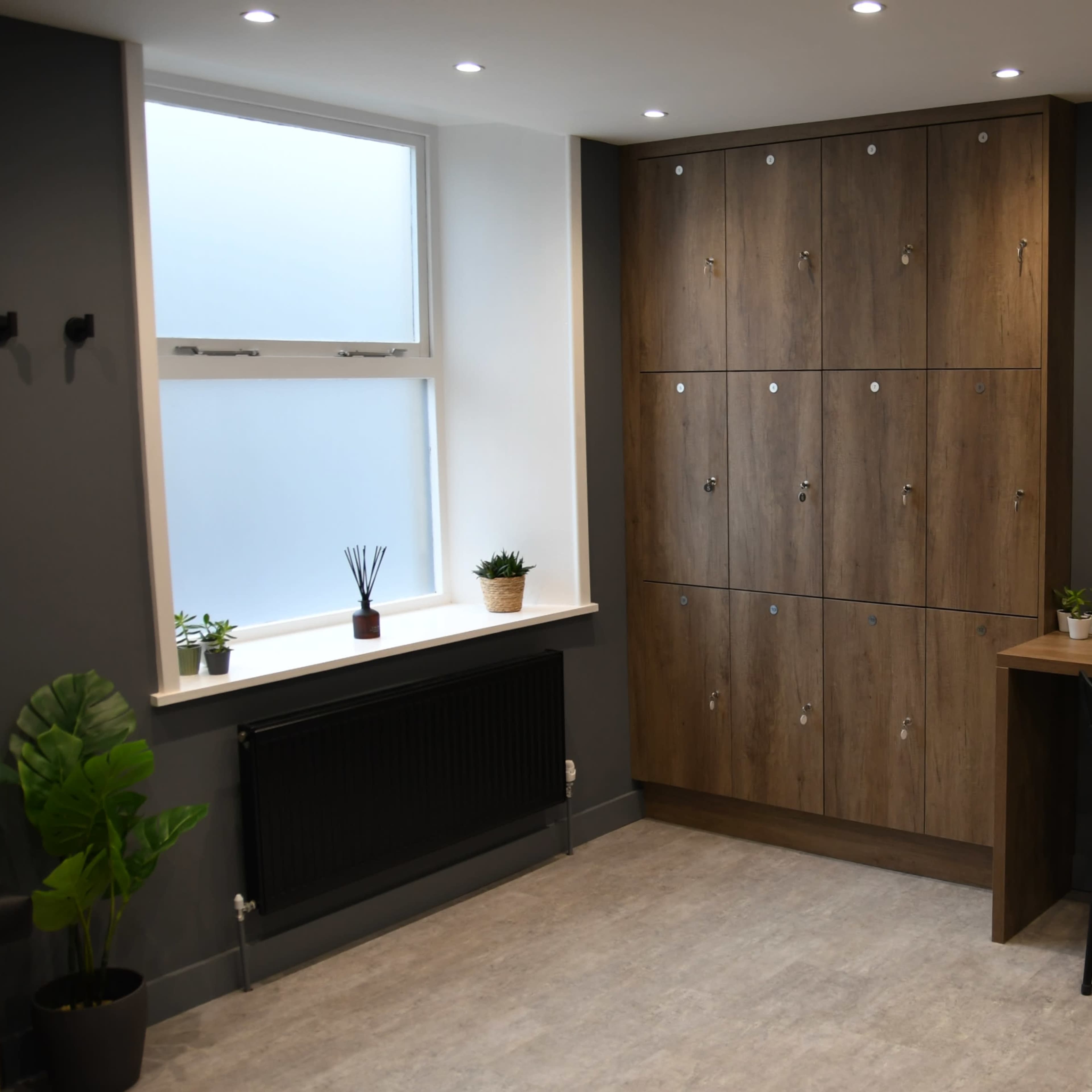 A modern room with gray walls, a wooden locker unit, two oval mirrors, a styled countertop with hairdryers, and potted plants.