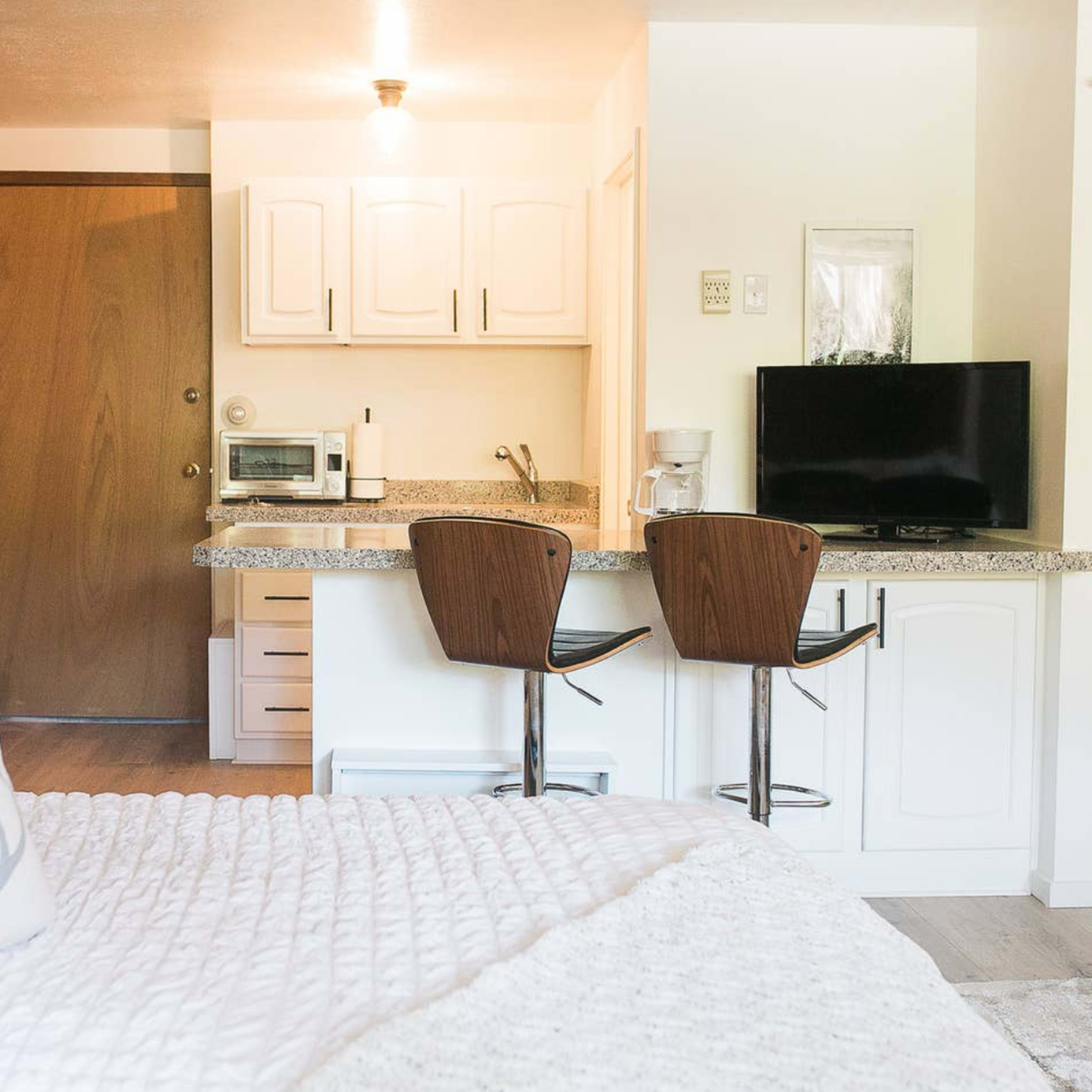 A cozy studio apartment with a bed in the foreground and a kitchen area featuring a bar with two stools and a television in the background.