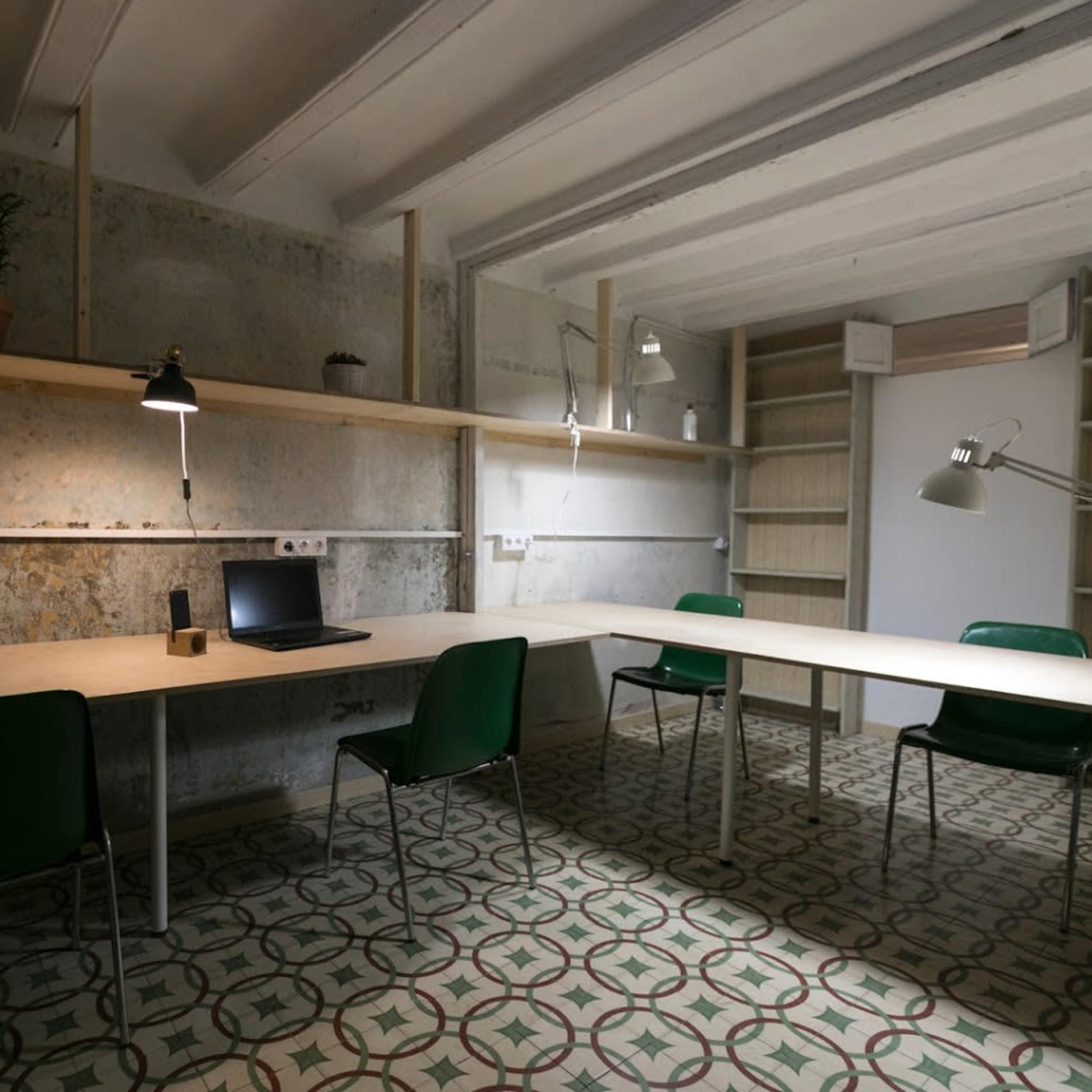 A minimalistic workspace with a large table, four green chairs, a laptop, and a potted plant against a textured wall.