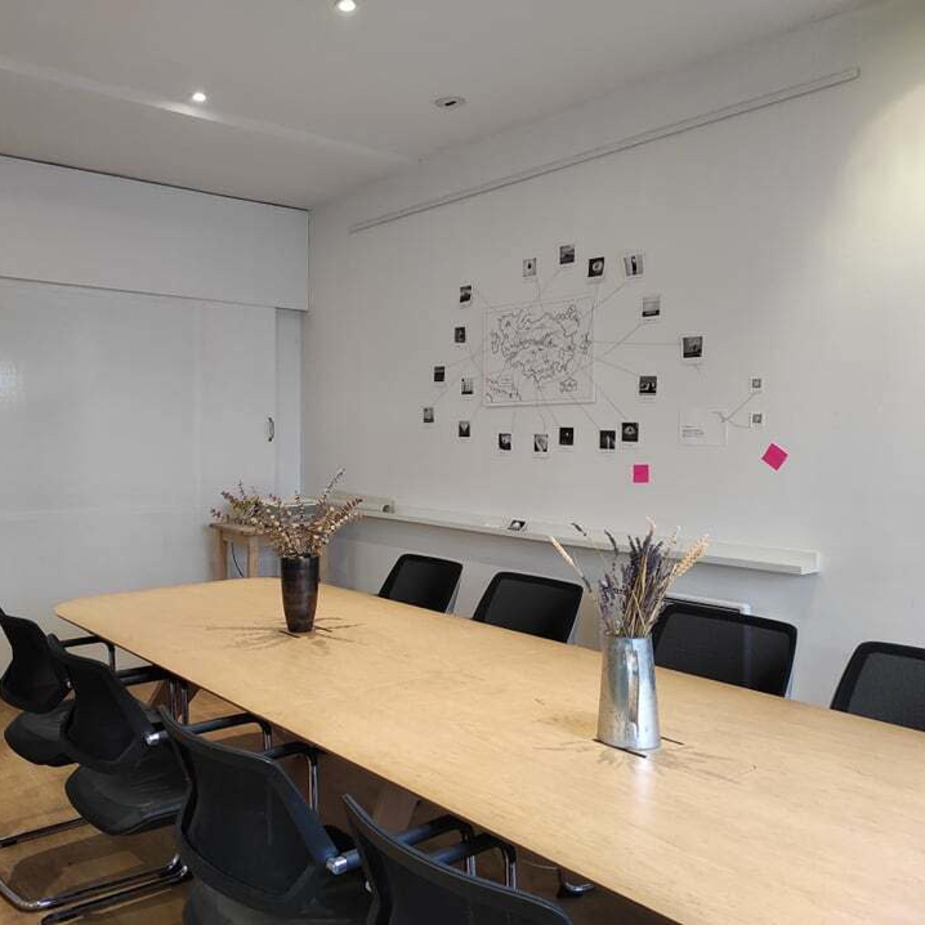 A long wooden table is set in a conference room with black chairs, a whiteboard, and various photos taped to the wall.