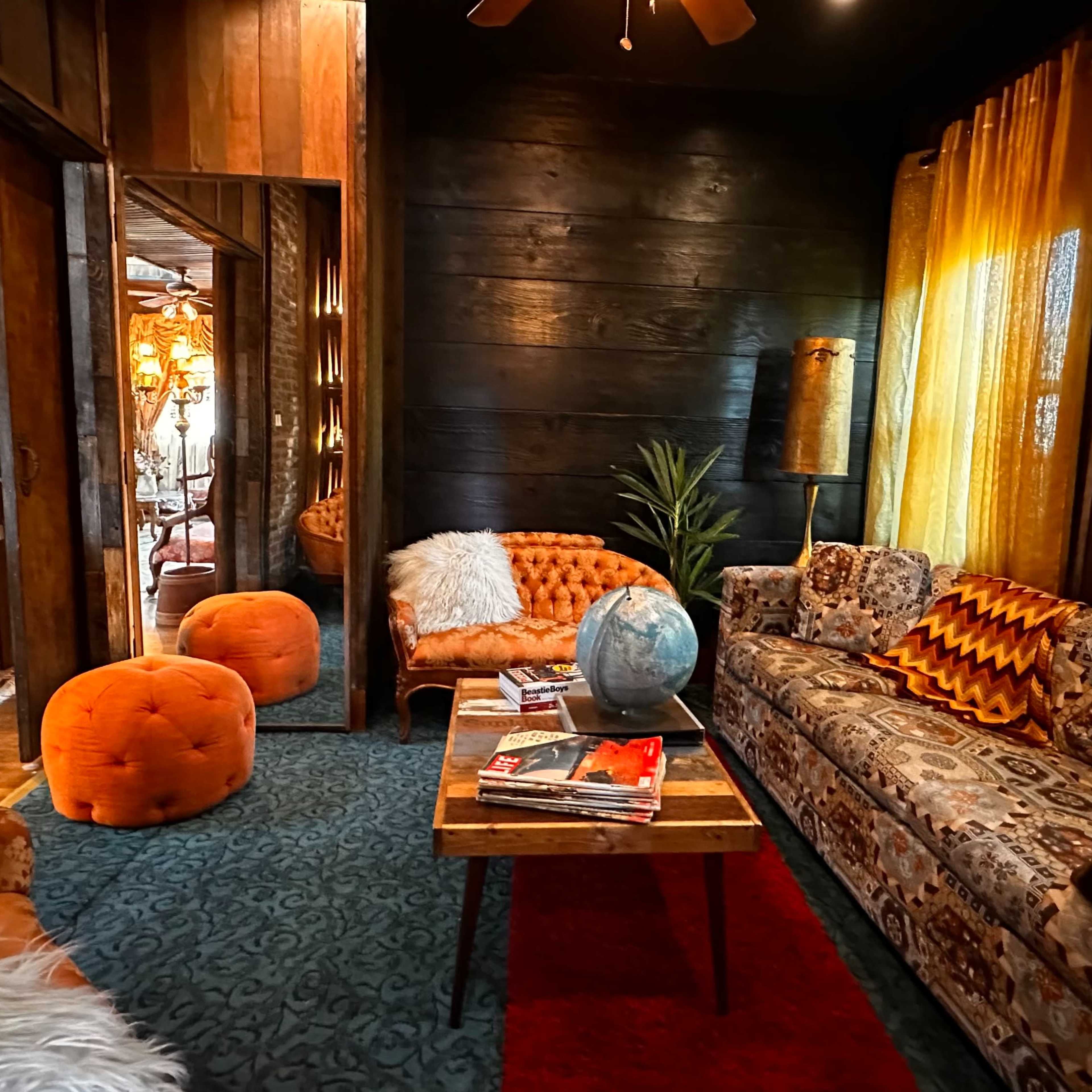 The cozy living area features a mix of patterned sofas, an orange pouf, a wooden coffee table with magazines, and a globe, all set against dark wooden walls and bright yellow curtains.