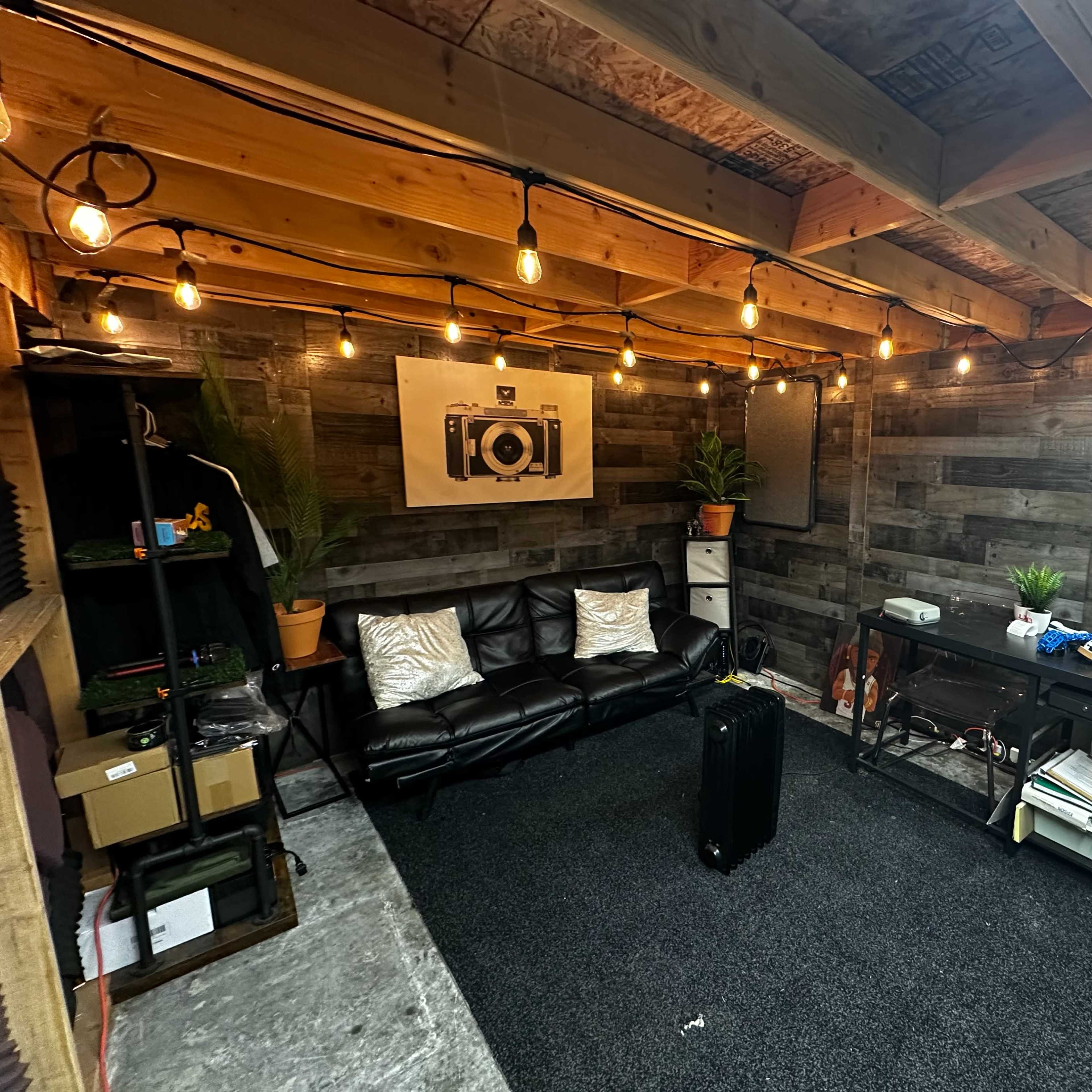 A well-lit interior space with wooden walls, string lights, a black sofa, and a small workspace equipped with various items and plants.