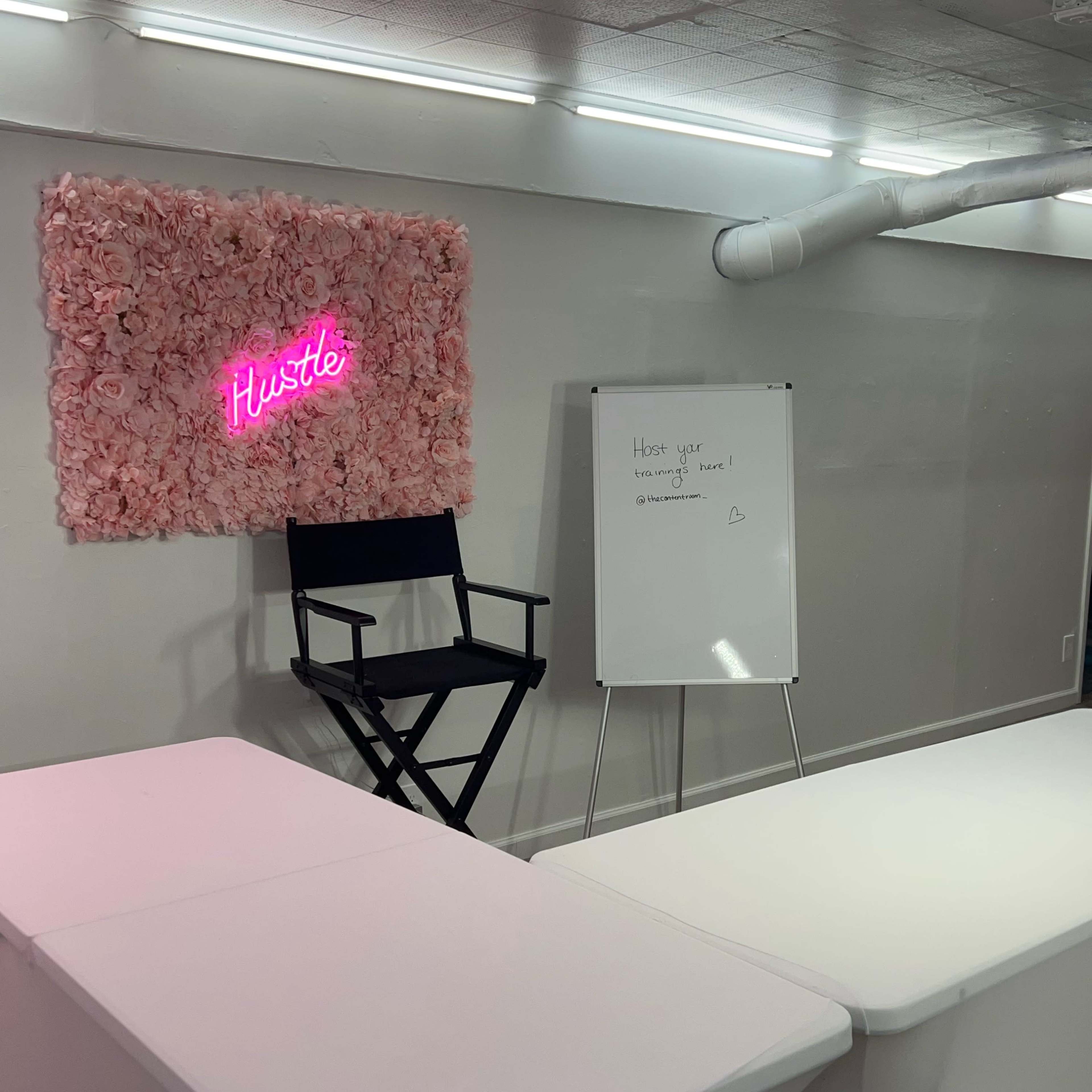 A minimalist workspace featuring a floral wall with a neon sign that reads "Hustle," a director's chair, and a whiteboard.