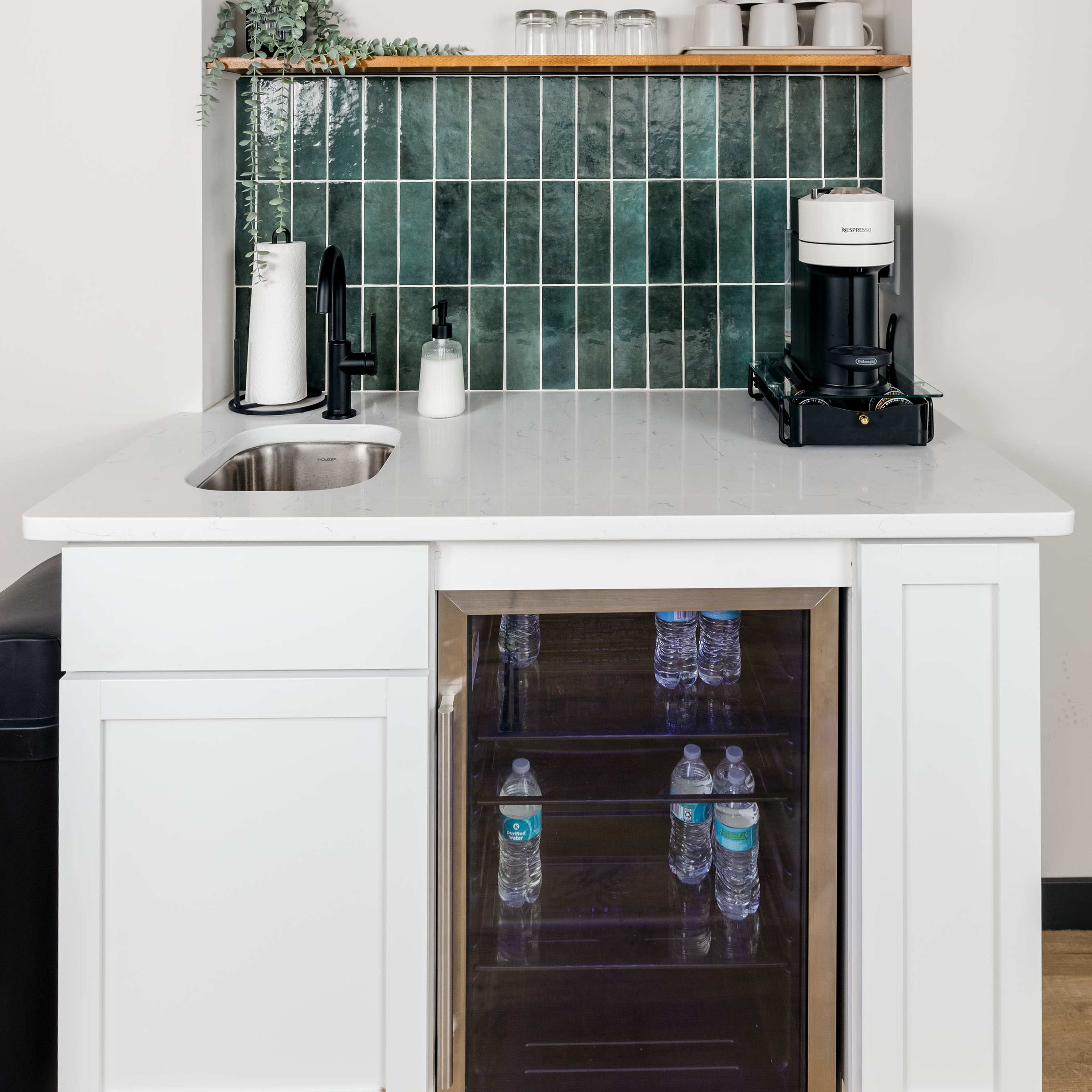 A modern kitchenette with a sink, a coffee maker, and a mini refrigerator displaying water bottles, set against a green tiled backsplash and wooden shelves.