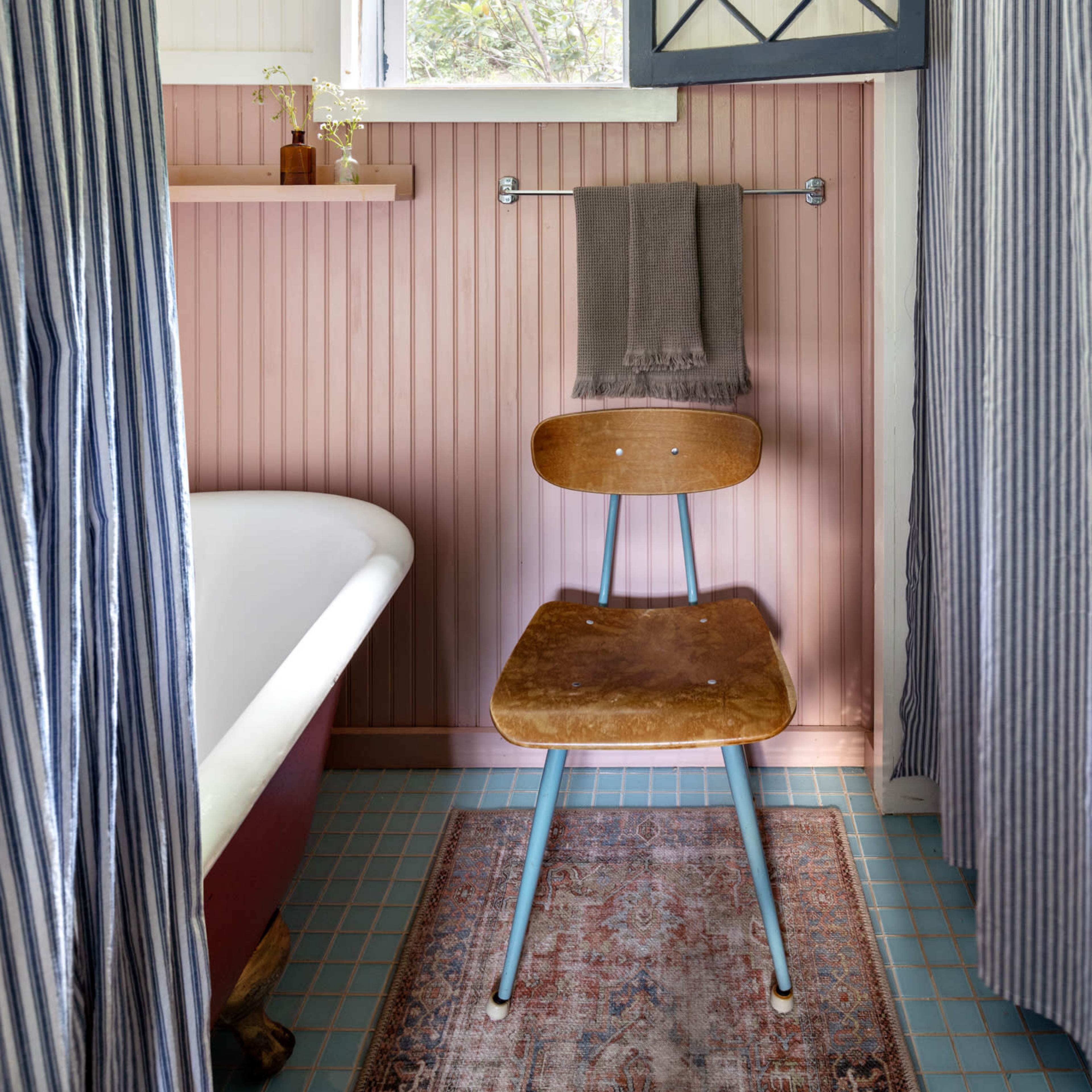 A bathroom with a clawfoot bathtub, a wooden chair, and striped curtains, along with a small window open to the outside.