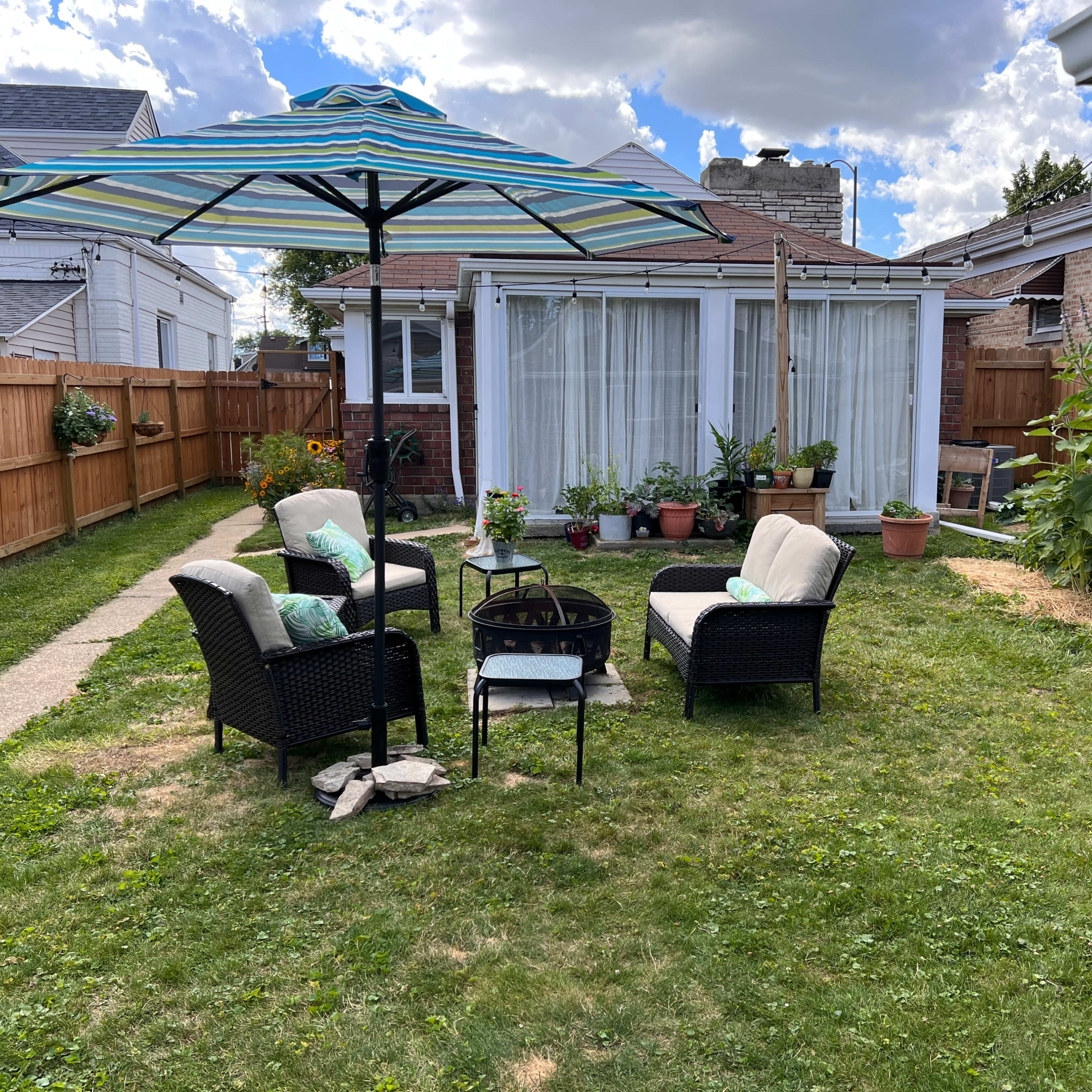 A backyard scene features a seating area with four chairs and an umbrella surrounding a fire pit, set against a backdrop of a garden and a house with a large window.