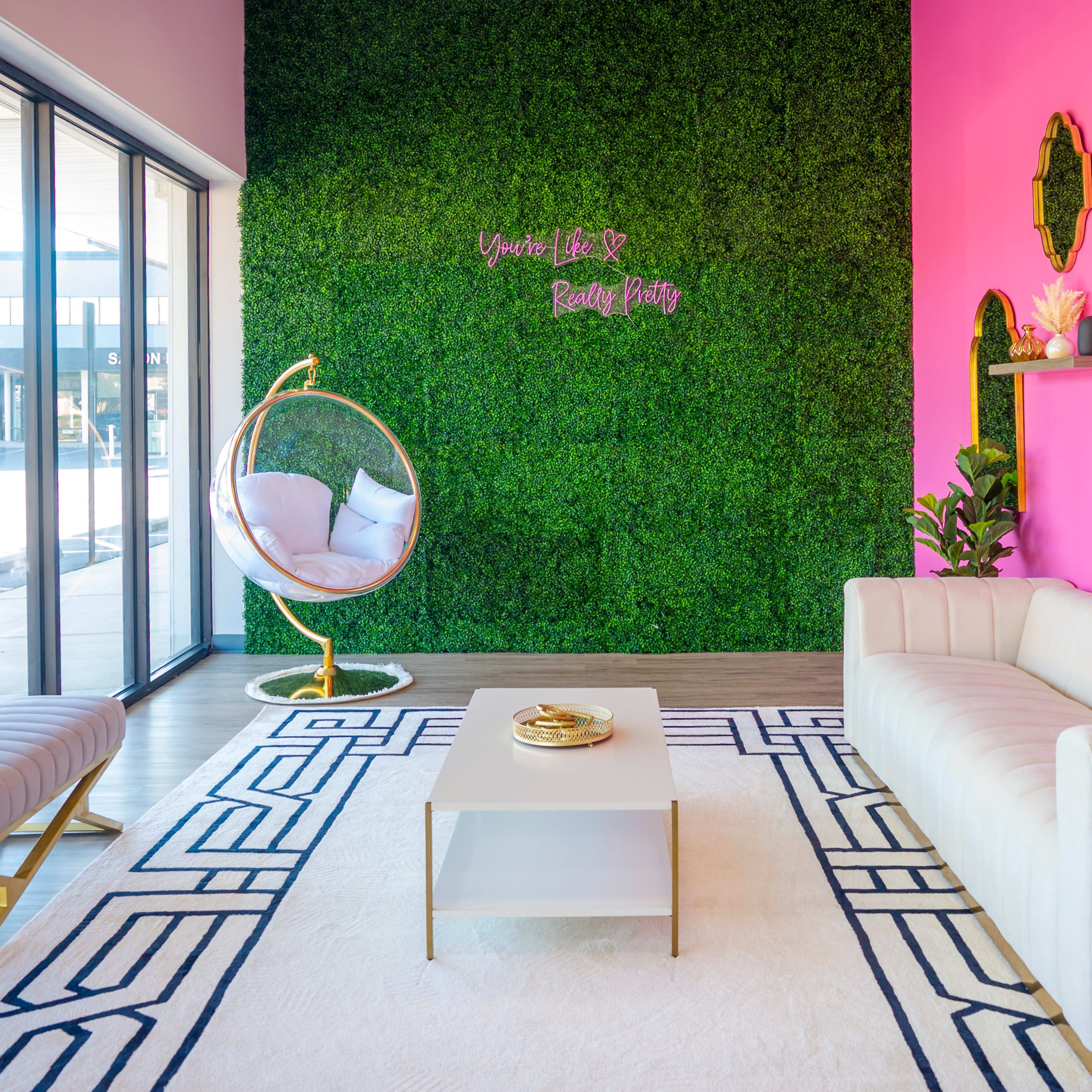 A brightly colored interior space with a pink accent wall, a green moss backdrop, a white sofa, a glass coffee table, and a hanging chair.