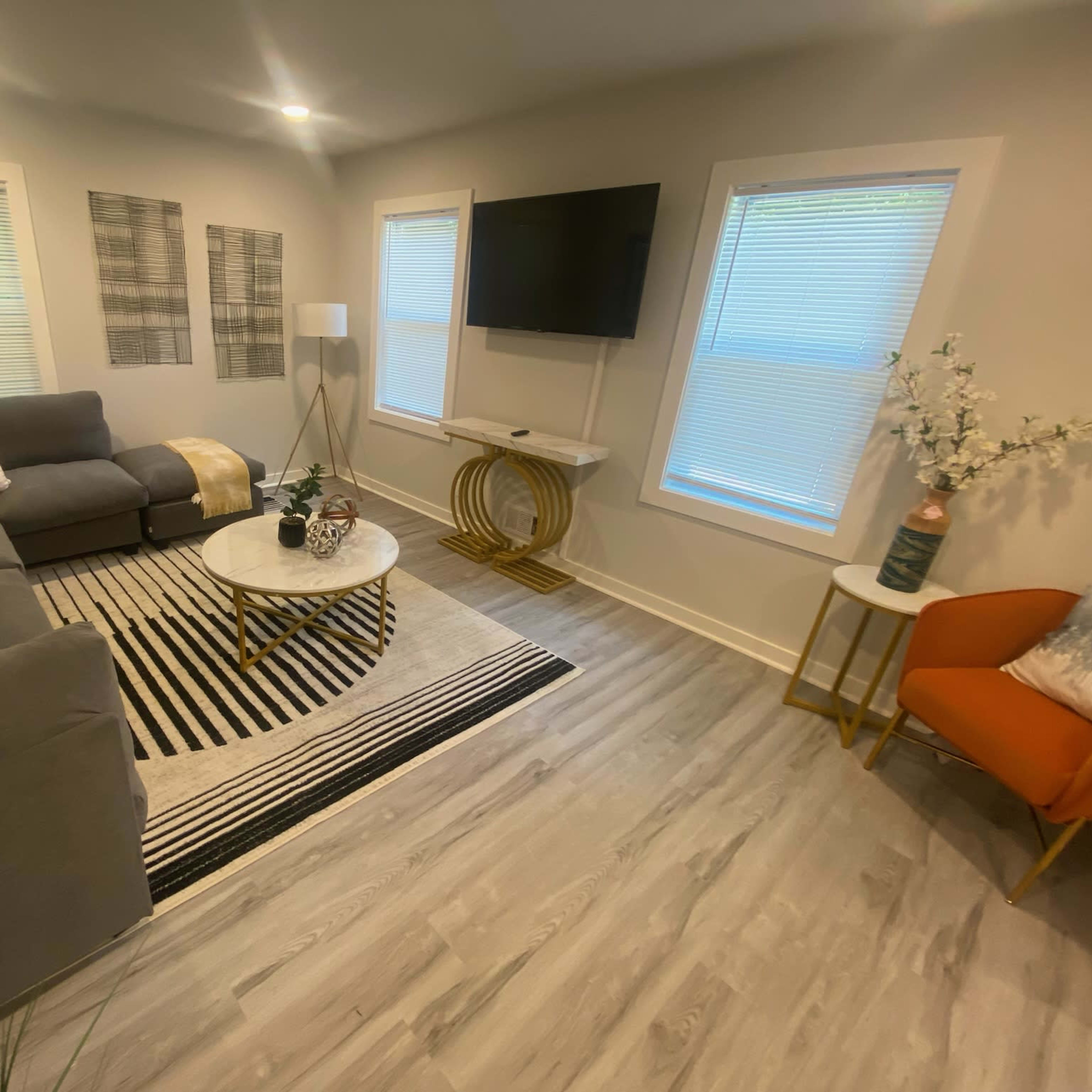A modern living room features a gray sectional sofa, a round coffee table, a striped area rug, and a television mounted on the wall under large windows.