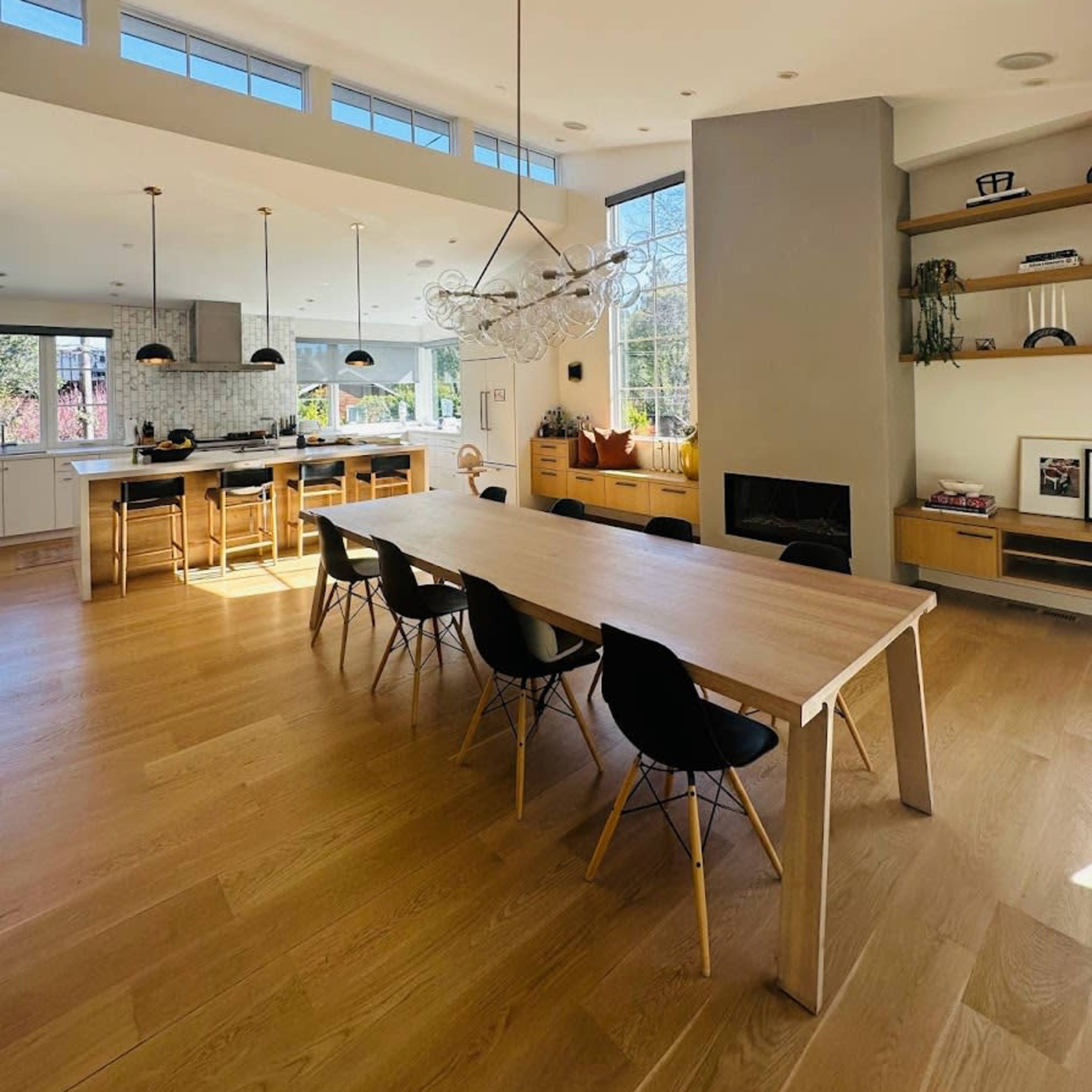 A modern kitchen and dining area features a long wooden table surrounded by black chairs, with large windows allowing natural light to fill the space.