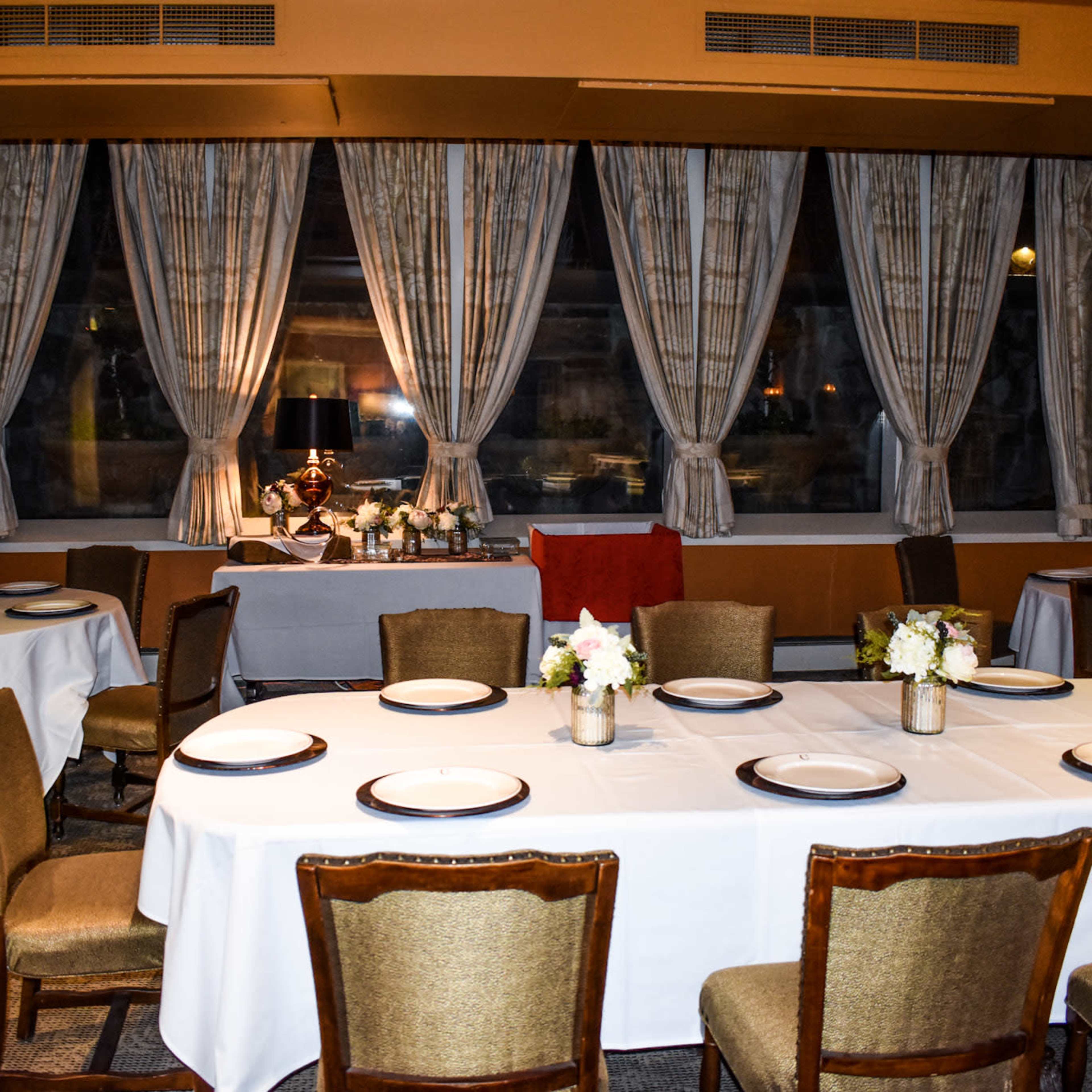 A dining room is set with white tablecloths and plates on round tables, facing windows draped with curtains.