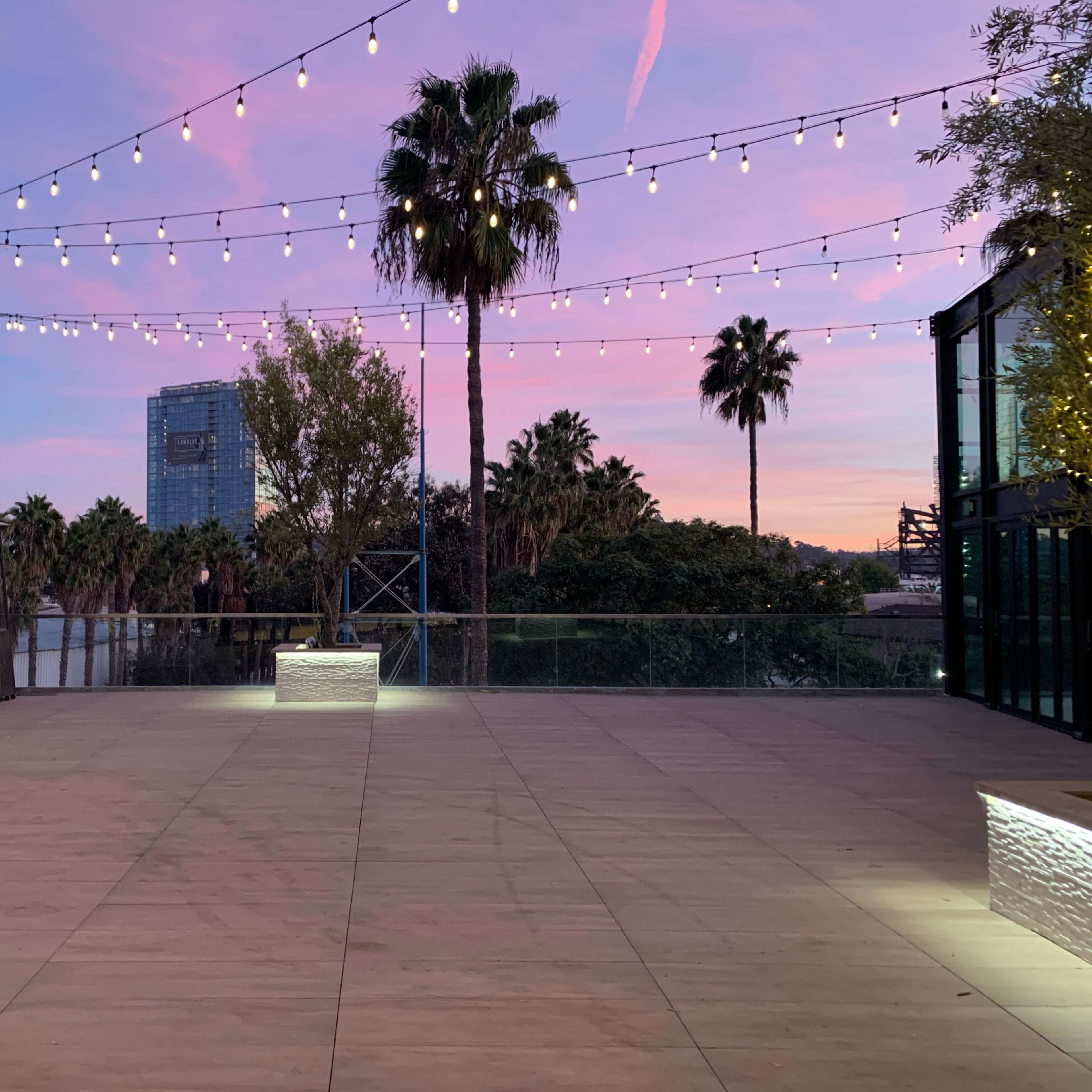 A spacious outdoor area with string lights overhead, palm trees in the background, and a colorful sunset sky.