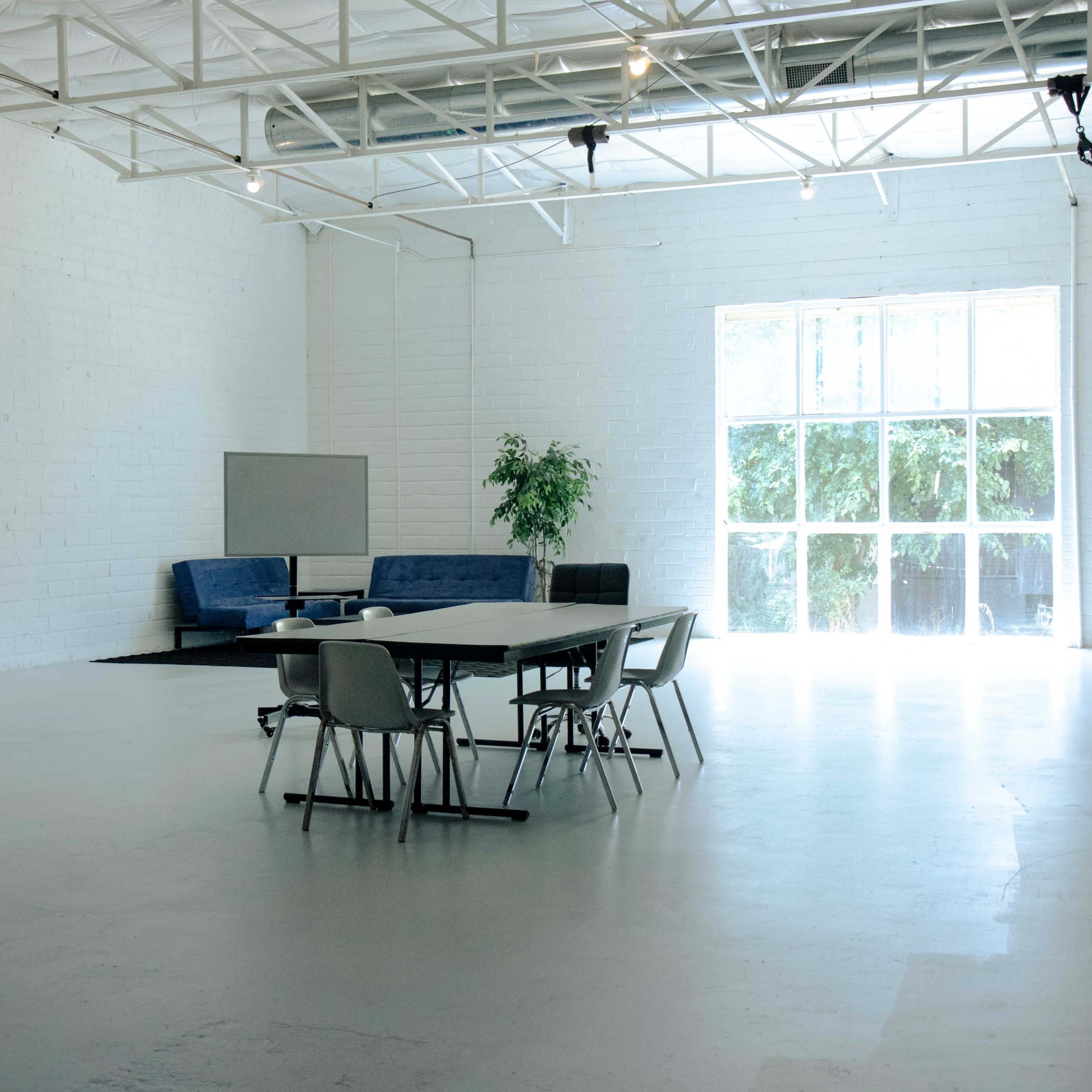 A spacious, minimalist room with a large table surrounded by chairs, a sofa in the corner, and a large window letting in natural light.