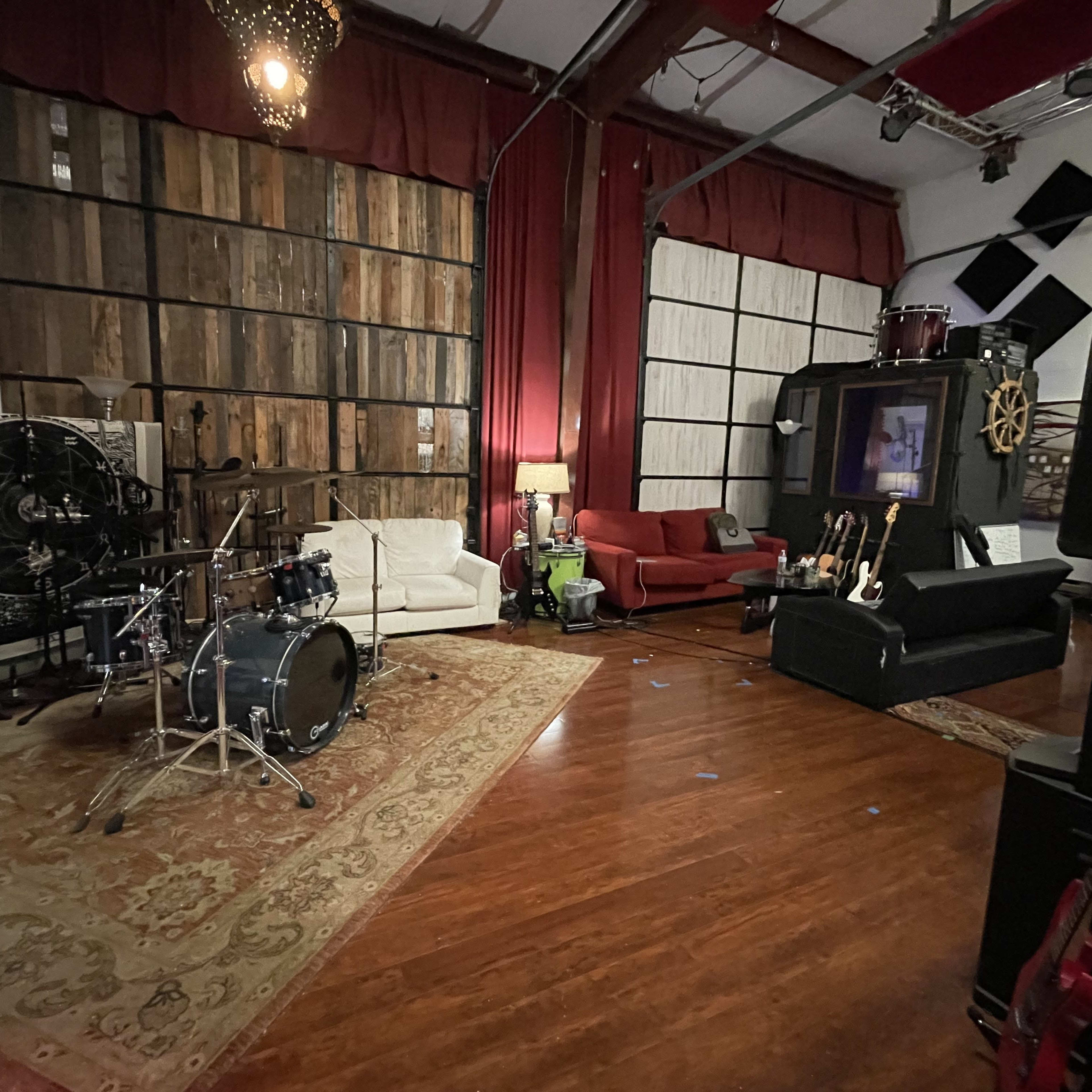 A music studio features a drum set, various guitars, and seating areas adorned with a large rug and decorative wall elements.