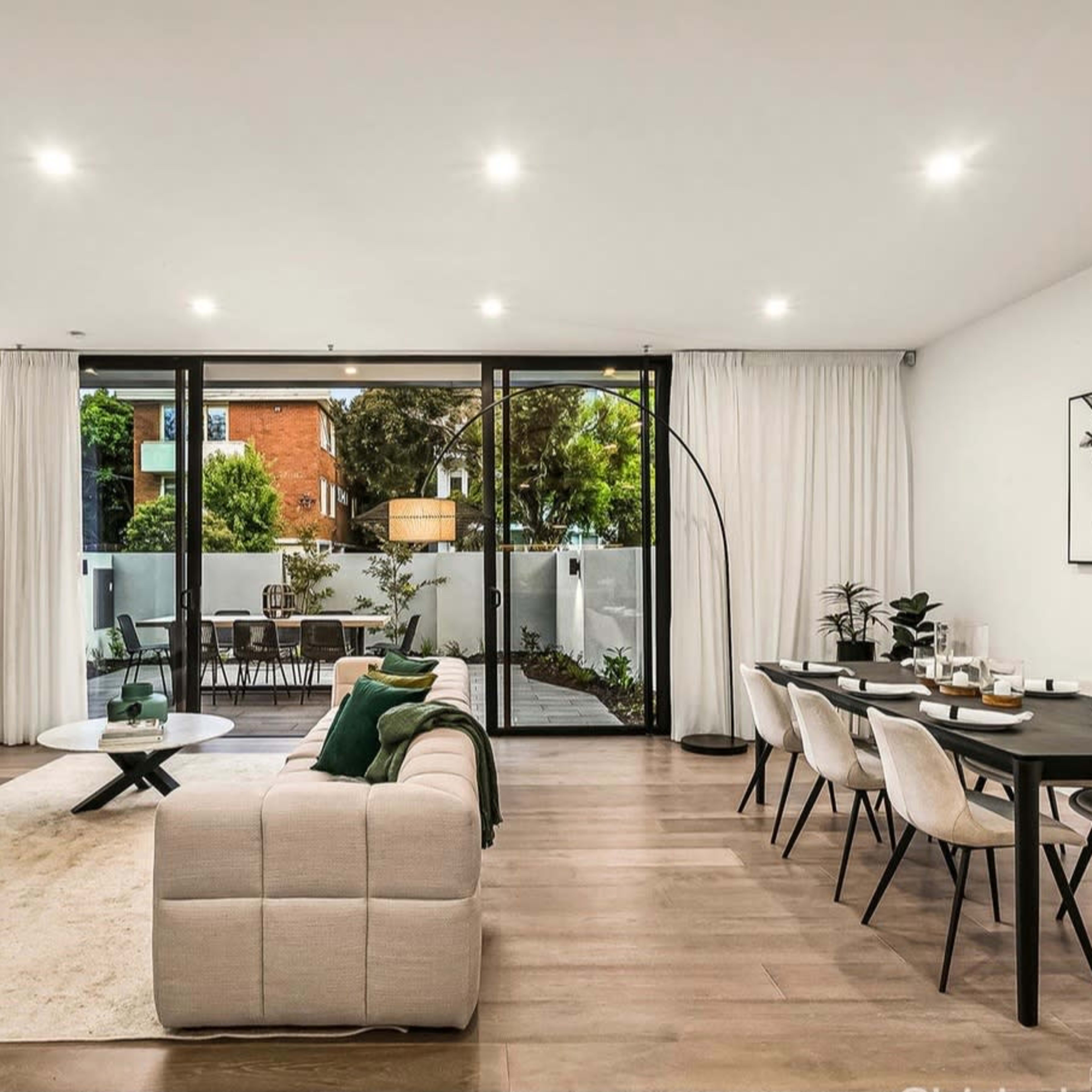 A modern living room features a sectional sofa, a coffee table, a dining area with a black table, and large glass doors opening to an outdoor space with greenery.