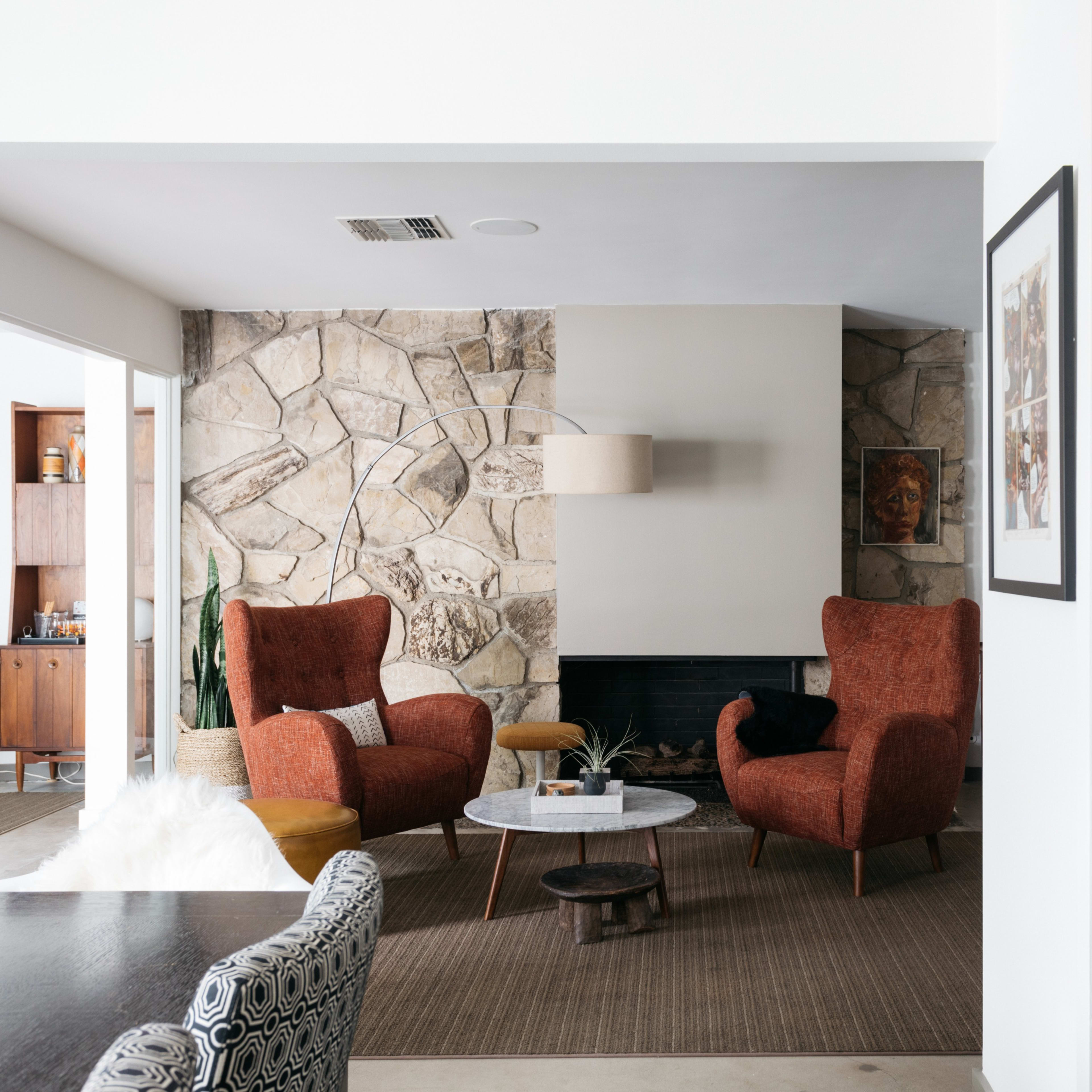A modern living room features two orange armchairs facing a stone wall, with a coffee table and a kitchen area visible in the background.