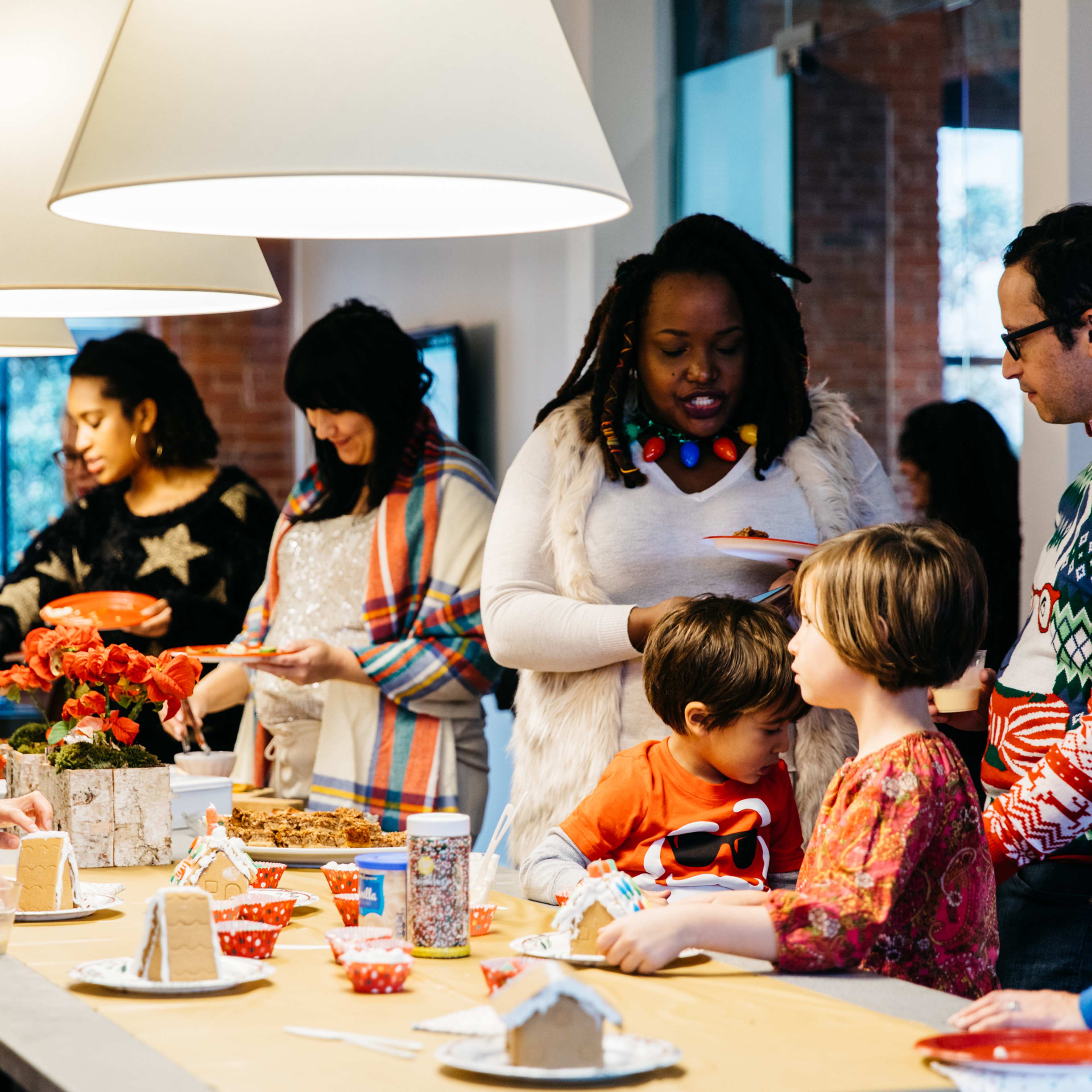 A group of people in festive sweaters gathers around a table decorated with holiday treats and crafts in a bright, open space.