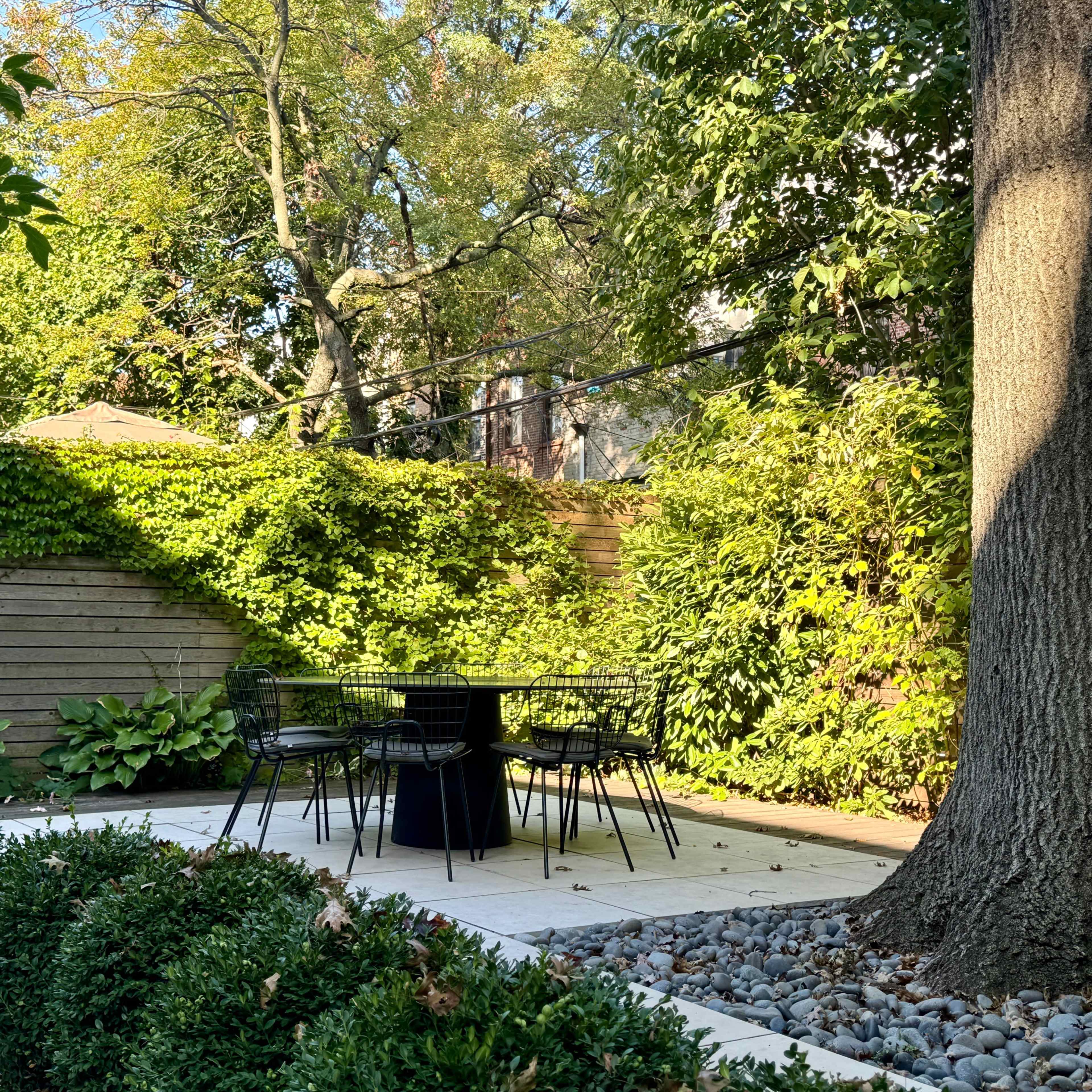 A small outdoor seating area features a black table and chairs surrounded by greenery and stone pathways.