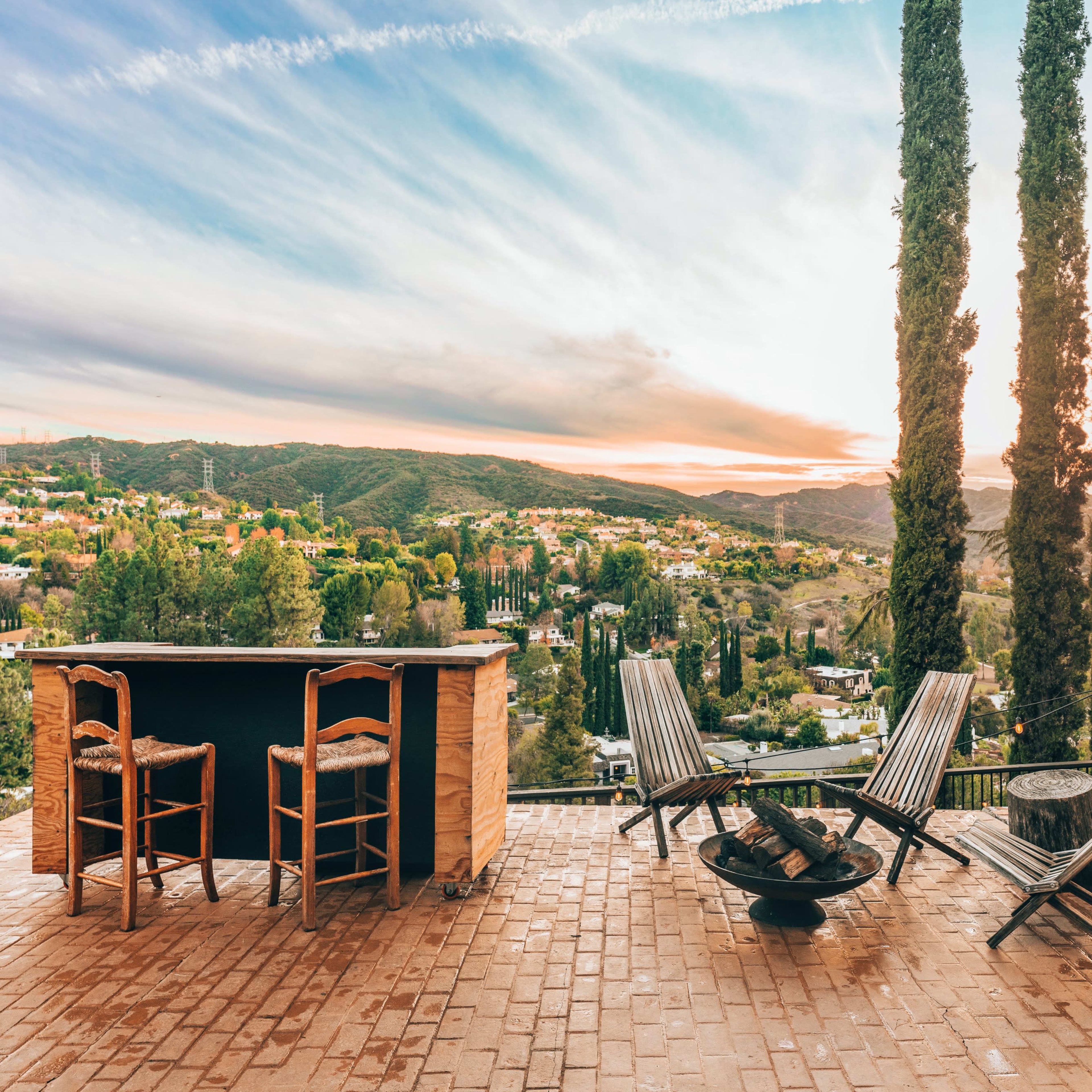 A patio with wooden chairs overlooks a mountainous landscape at sunset, framed by tall cypress trees.