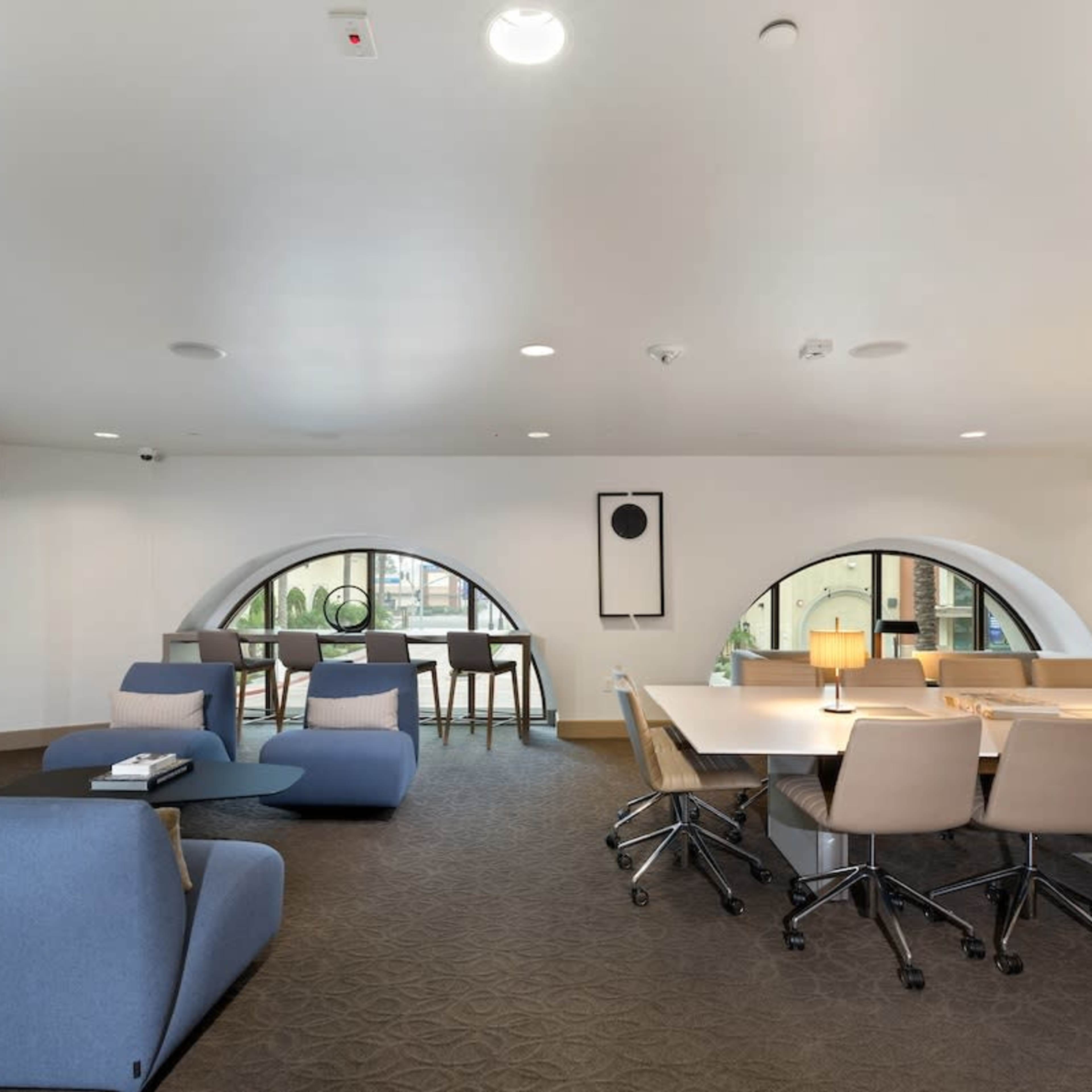 An office space with curved windows, a seating area with blue chairs, and a long table surrounded by light-colored chairs.