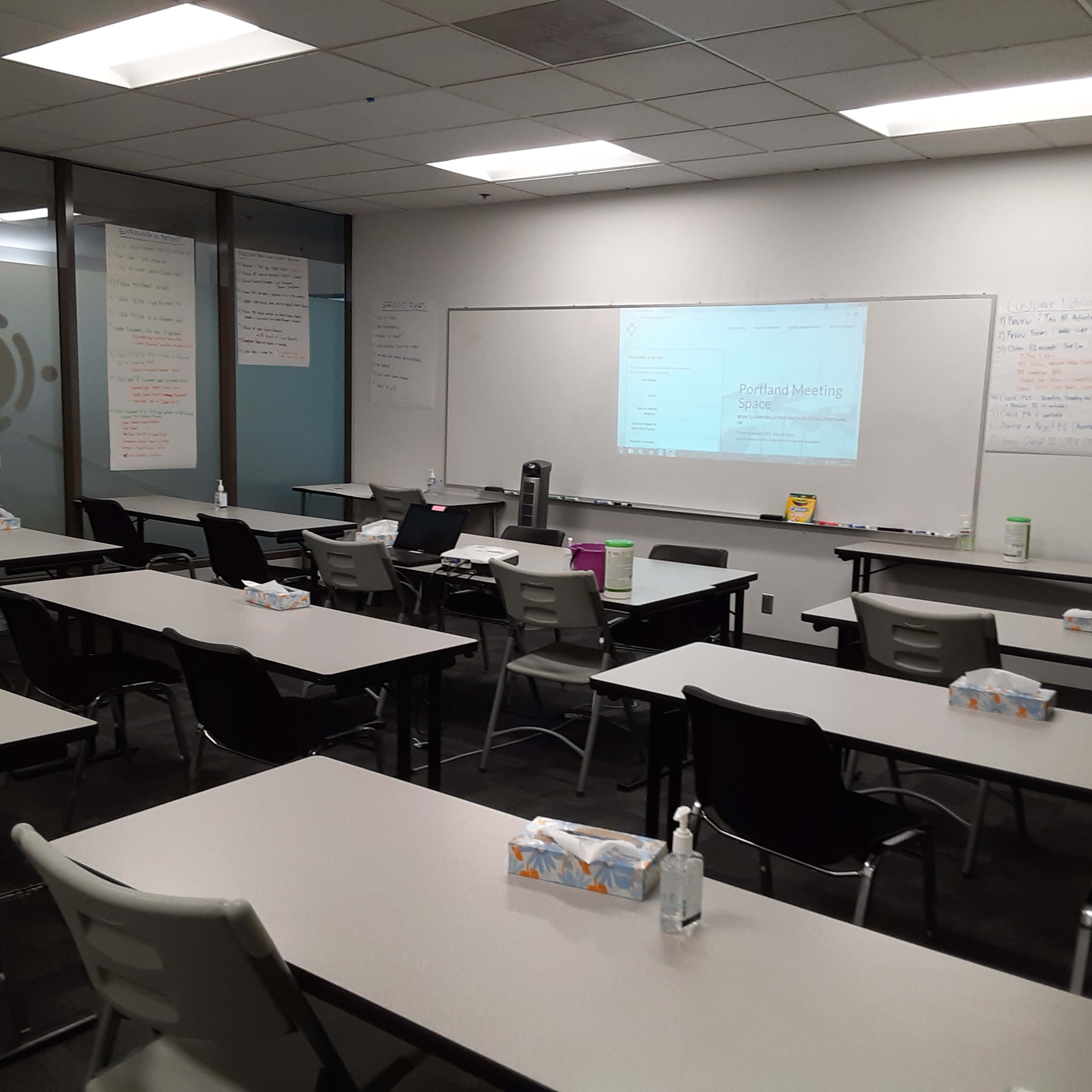 A classroom is set up with multiple tables and chairs arranged for a meeting, featuring flip charts and a whiteboard in the background.