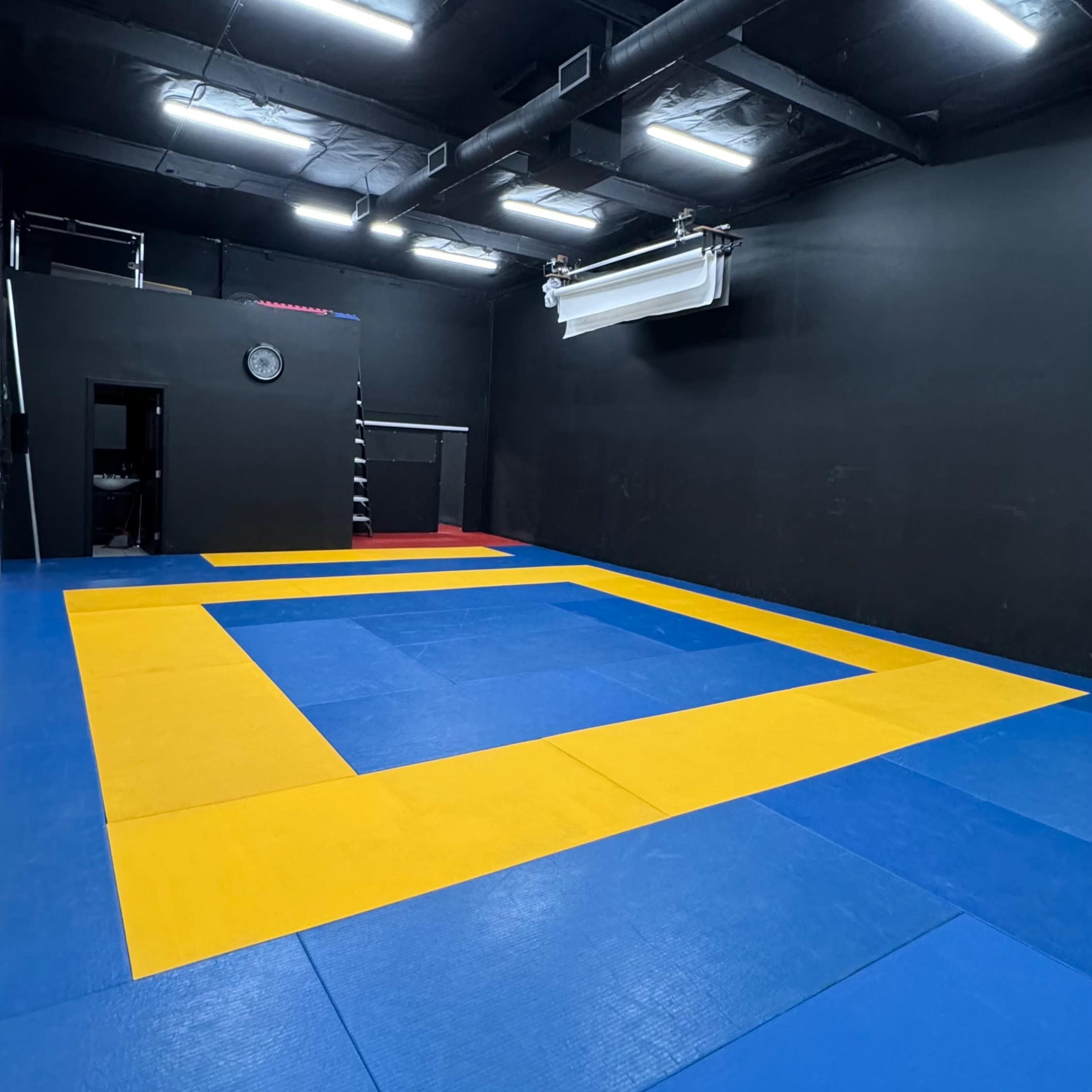 A sparsely furnished training room with blue and yellow interlocking mats, black walls, and overhead lighting.