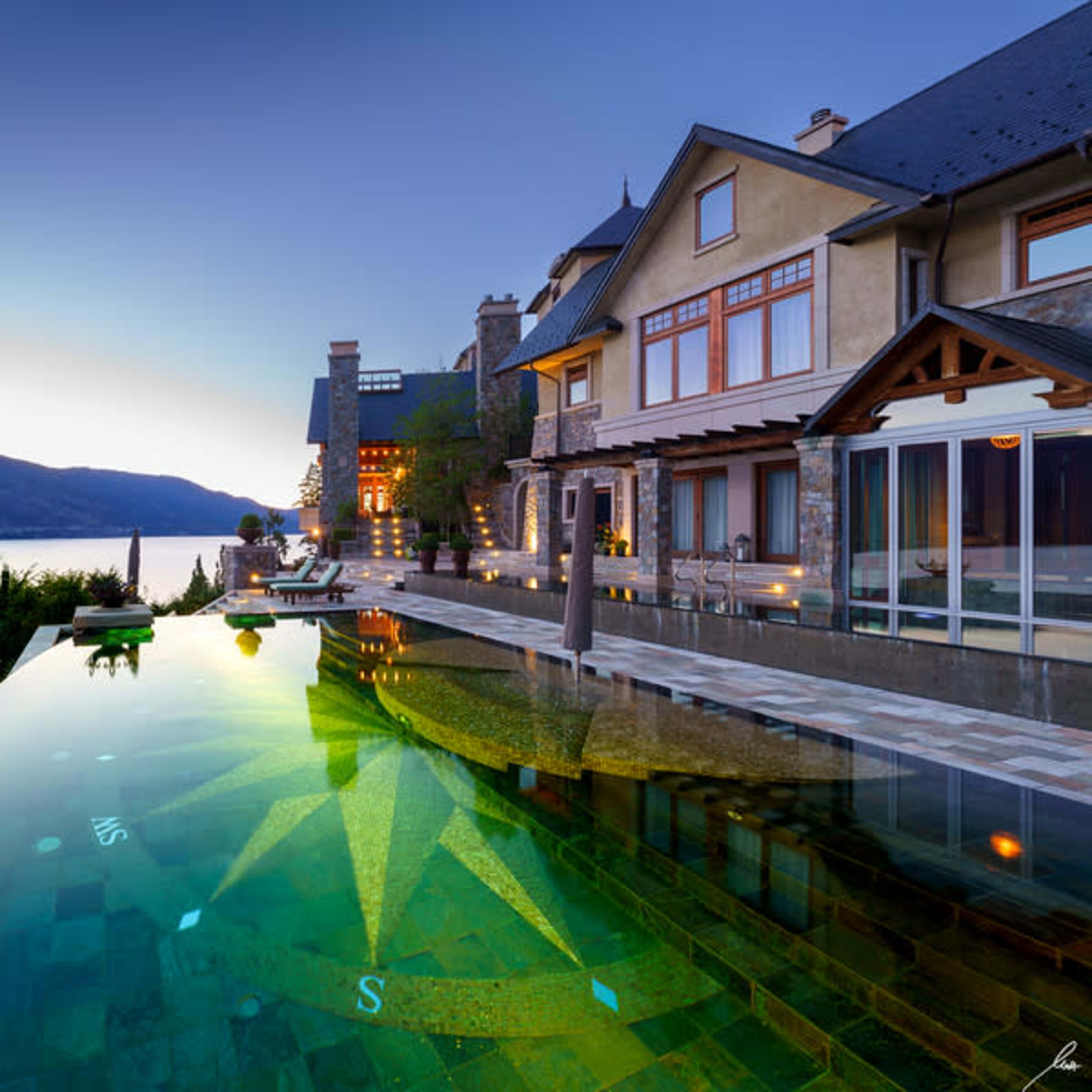 A large, luxurious home beside a tranquil water body, featuring a beautifully illuminated infinity pool with a star design at the bottom.