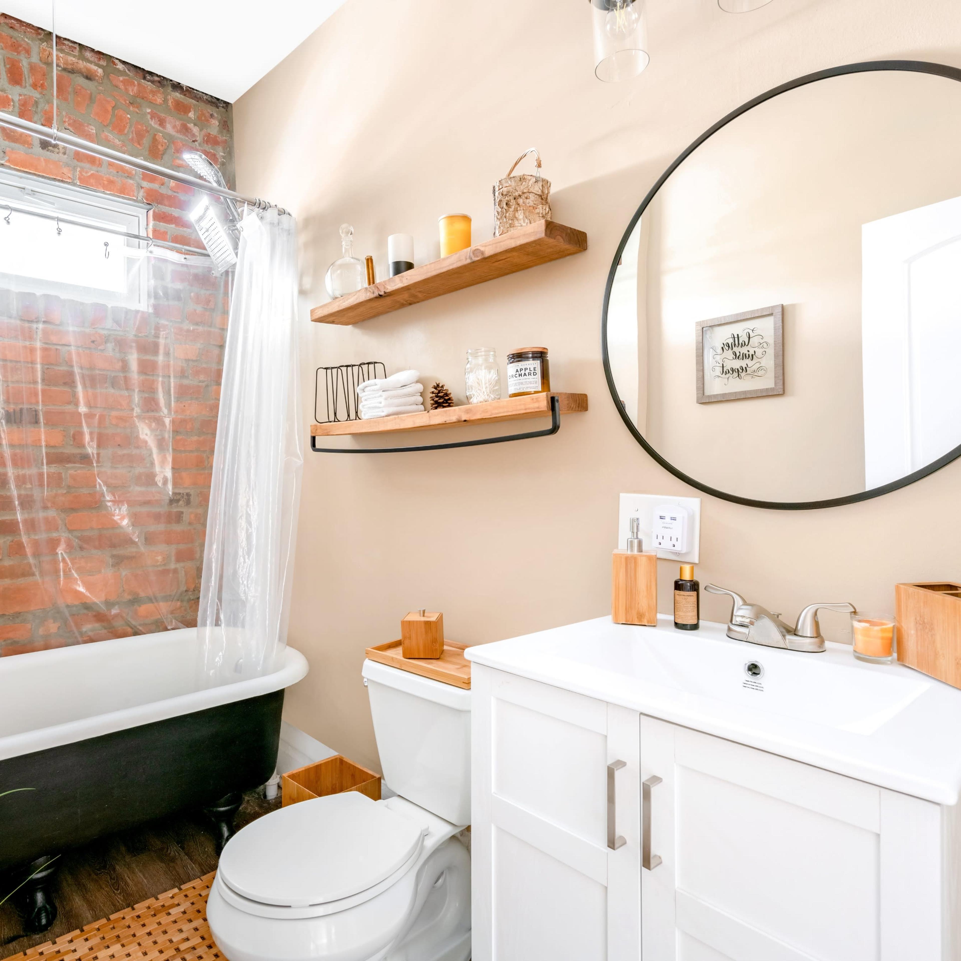 A bathroom featuring a black freestanding bathtub, a round mirror above a white vanity, and walls adorned with exposed brick and wooden shelves.