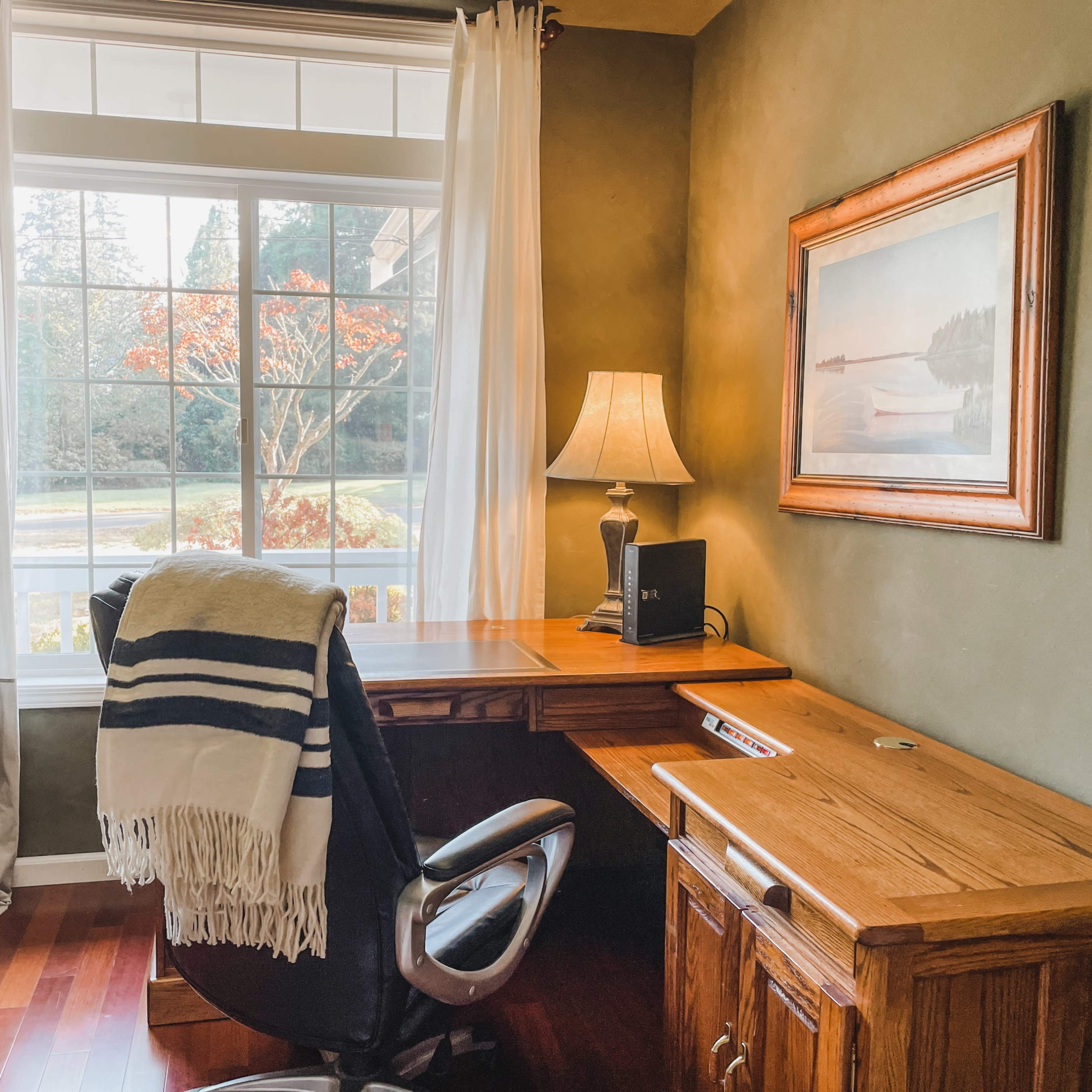A wooden desk with a chair is positioned near a window, which is adorned with curtains, and a lamp is placed on the desk alongside a painting on the wall.